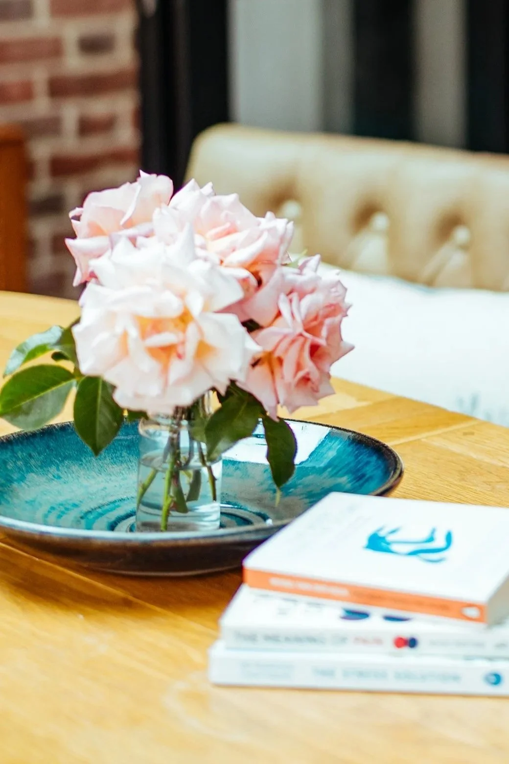 A small glass vase with pink and white flowers on a wooden table, with a blue ceramic dish underneath, and a white book with a blue logo beside the vase.