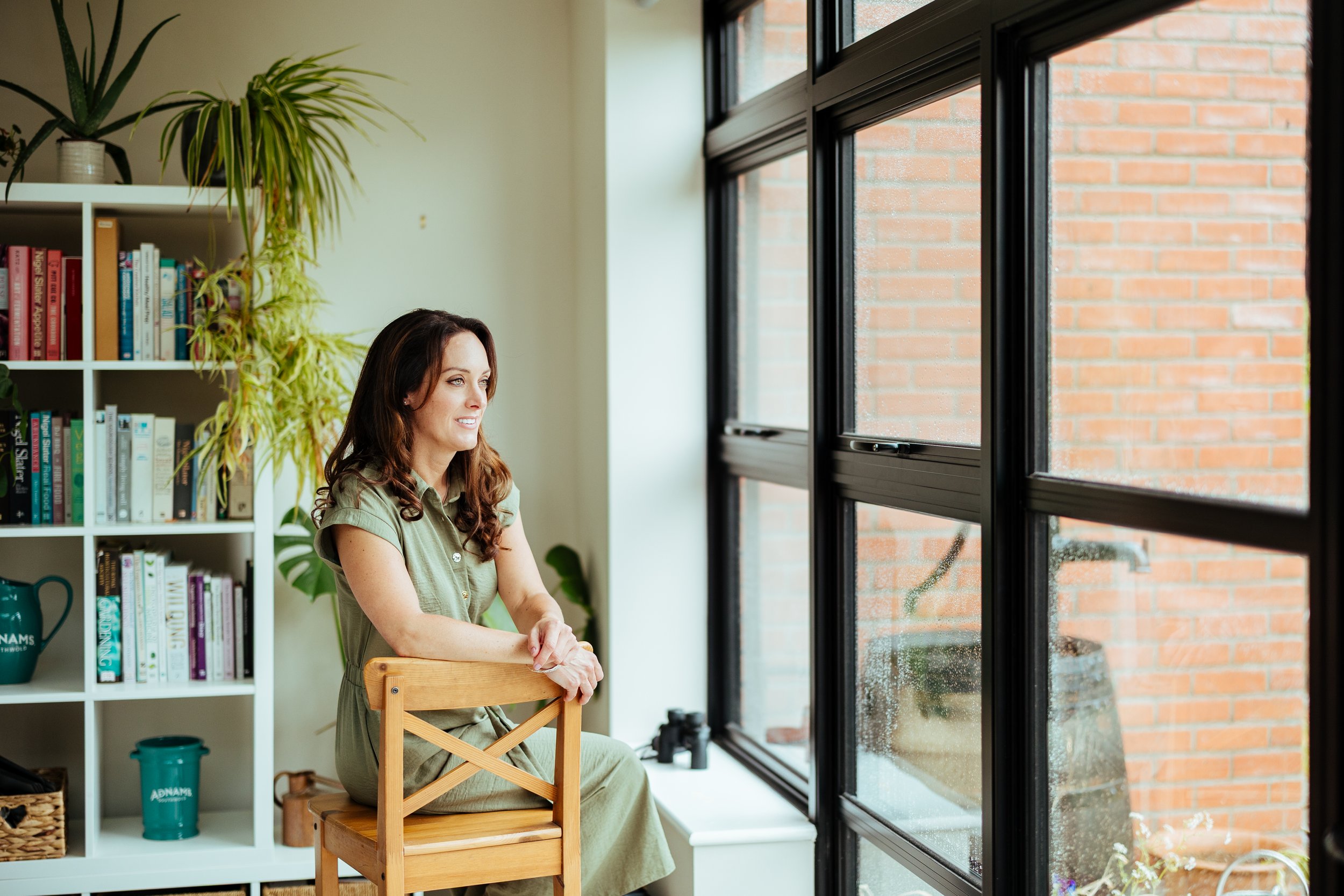 A woman sitting on a wooden chair by a large window, looking outside. She has long brown hair and is wearing a green dress. The room features a bookshelf with various books and houseplants.