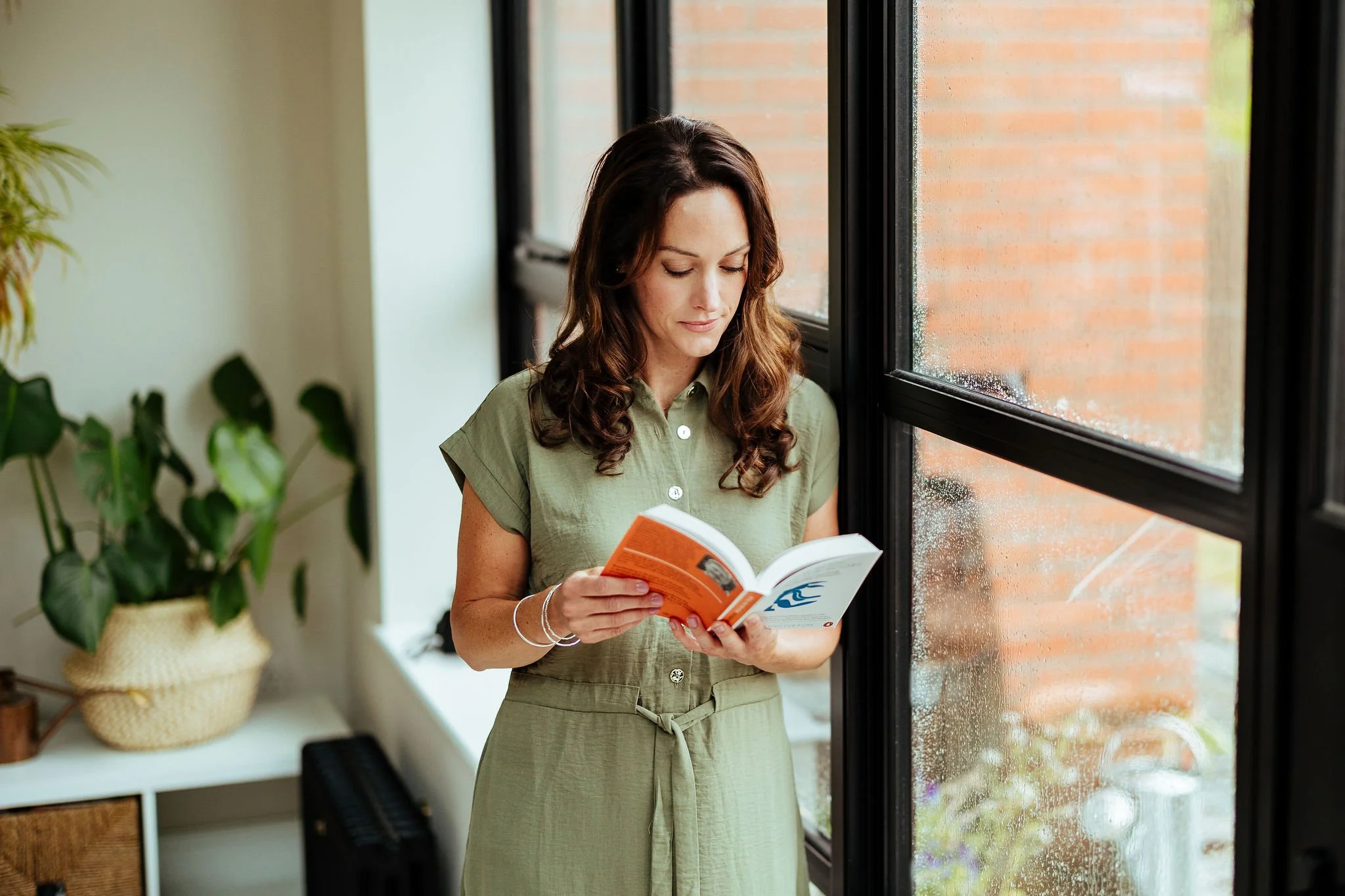 A woman with wavy brown hair wearing a light green dress reads a book while standing by a window on a rainy day.