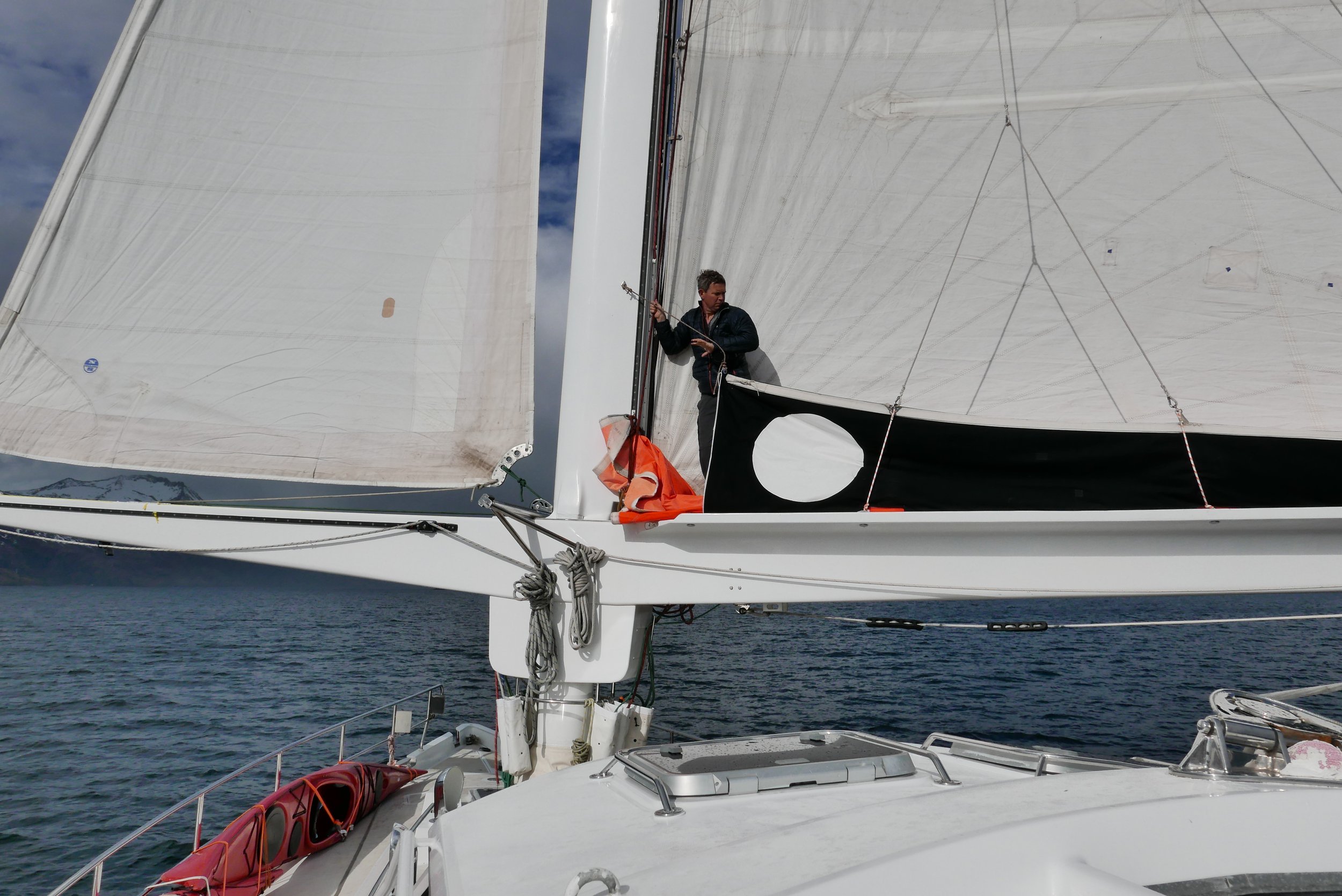 A person adjusting sails on a sailboat with white sails, black flag with a white circle, and a view of the ocean and cloudy sky in the background.