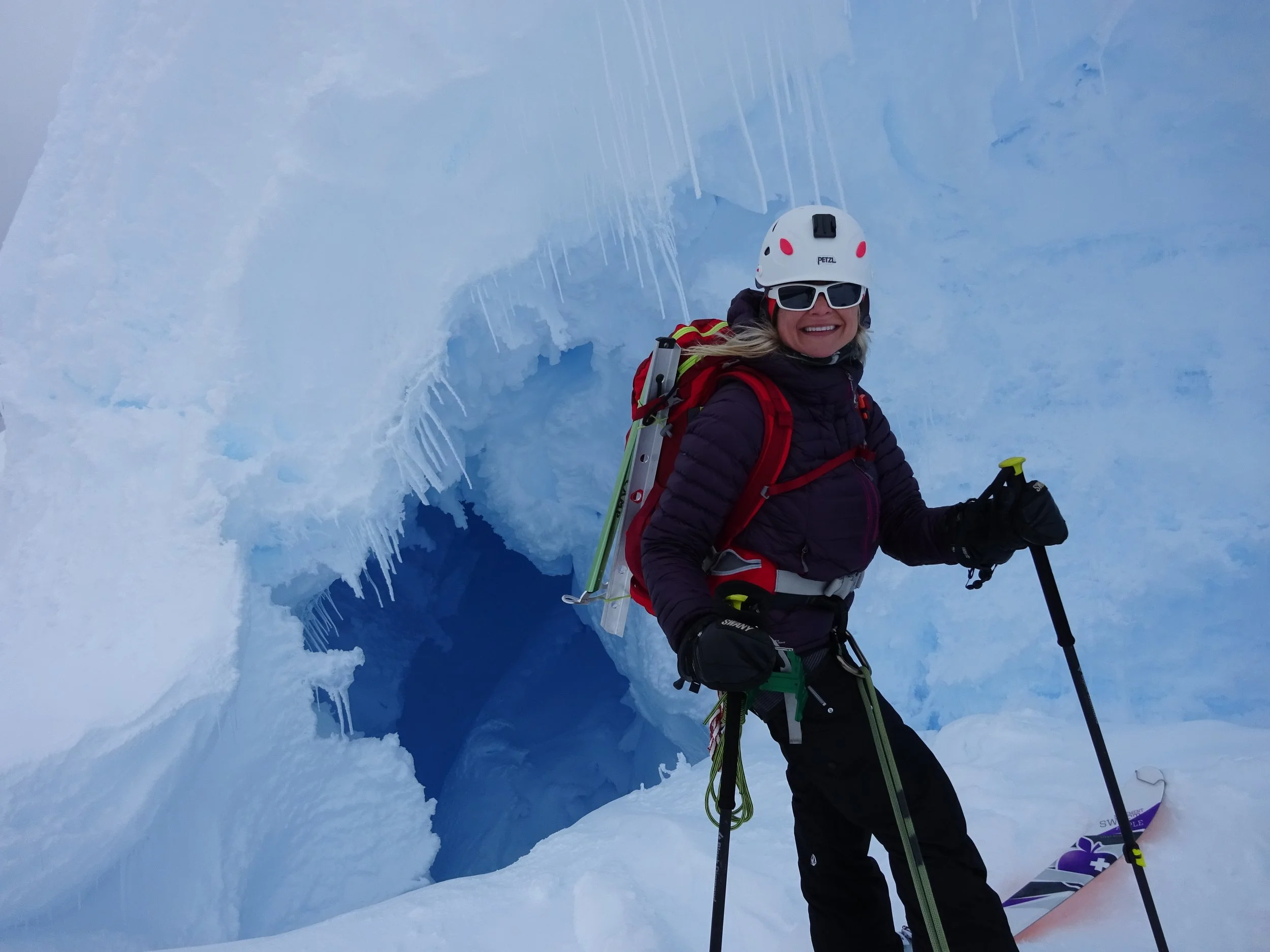 A woman in climbing gear standing in front of an ice cave with icicles, smiling and holding an ice axe and ski poles.
