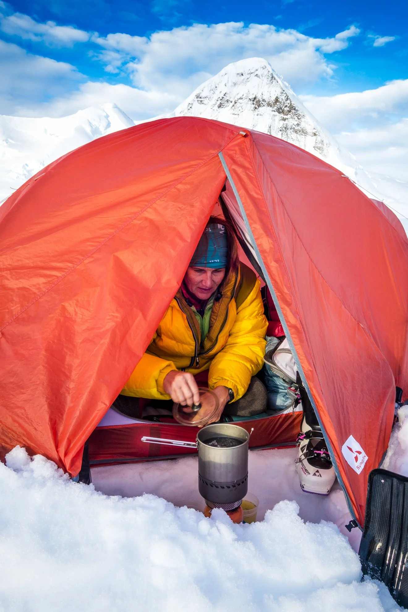 A person inside a red tent on snow with a snowy mountain in the background, preparing a hot drink using a portable stove.