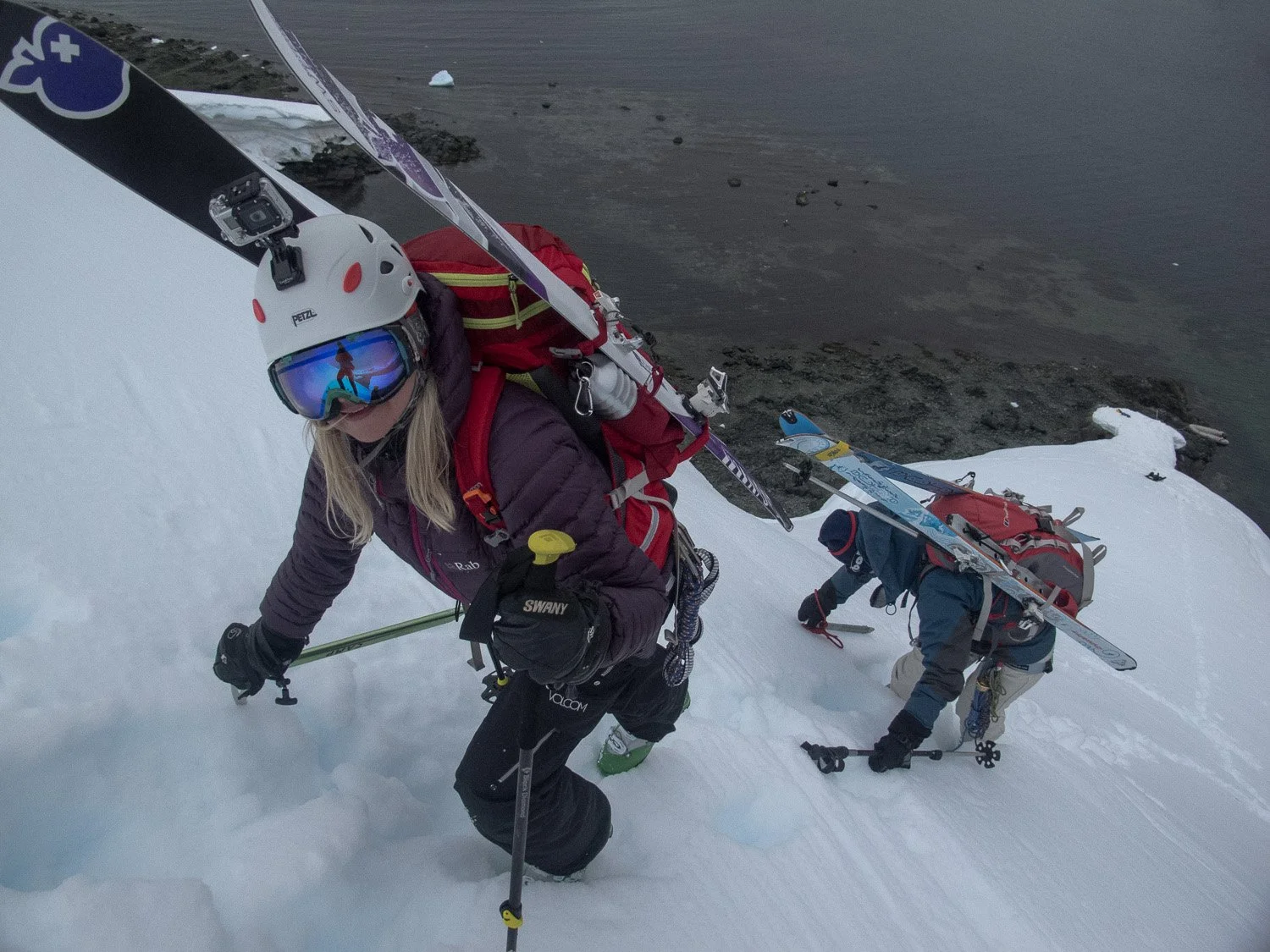Two people climbing a snowy mountain with gear, one in the foreground wearing goggles and a helmet, the other in the background, both using ice axes.