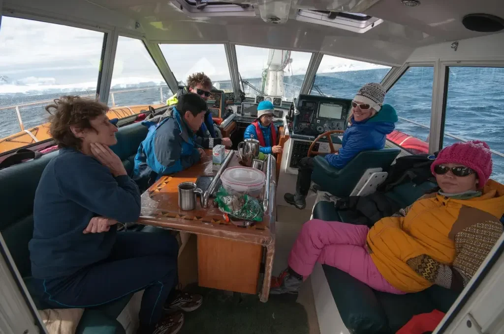 A group of six people on a boat, seated around a table and in the cockpit, with ocean visible outside.