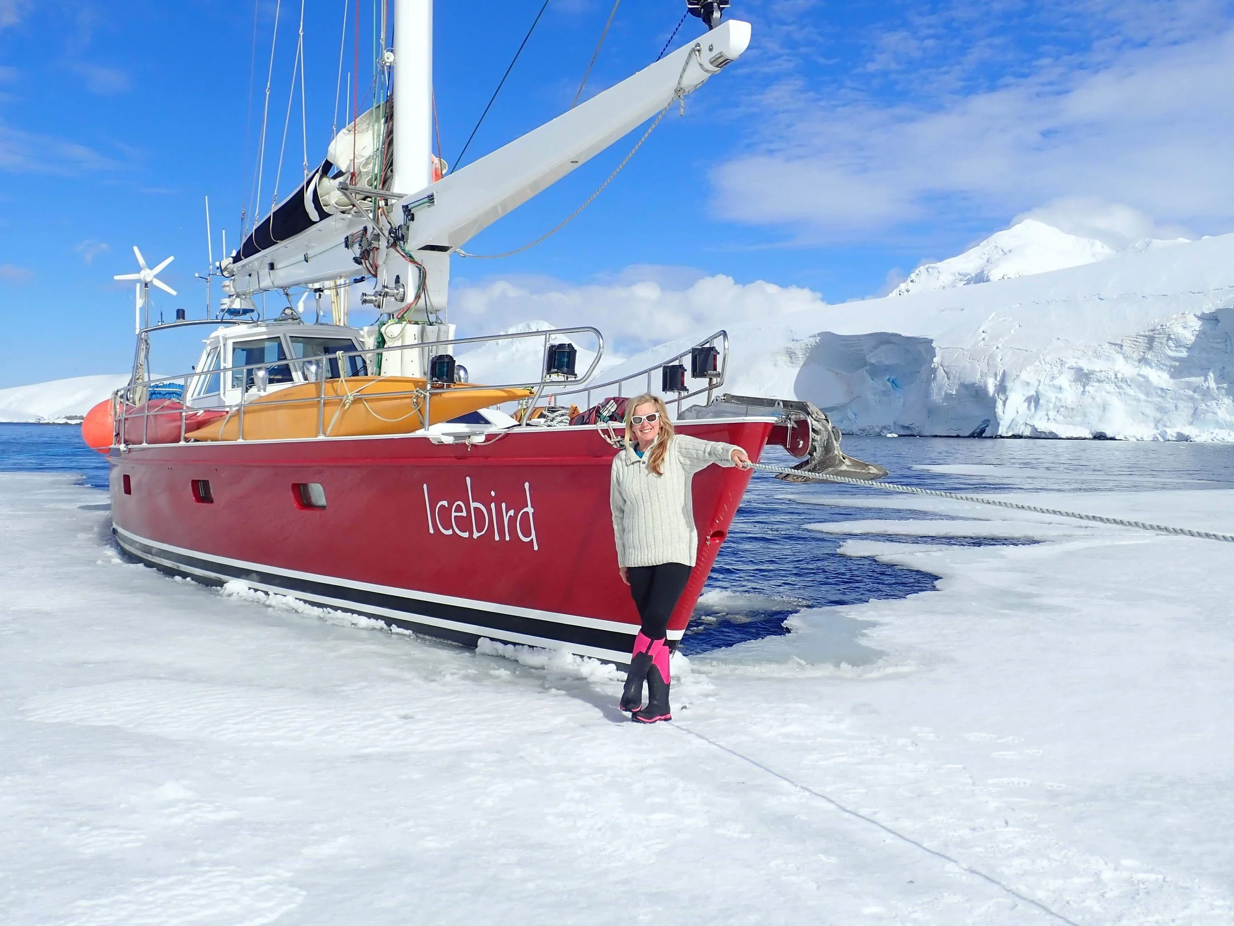 A woman in a white sweater and sunglasses stands on snow and ice, holding a rope attached to a red boat named 'Icebird' surrounded by icy landscape with snow-covered mountains and blue sky.