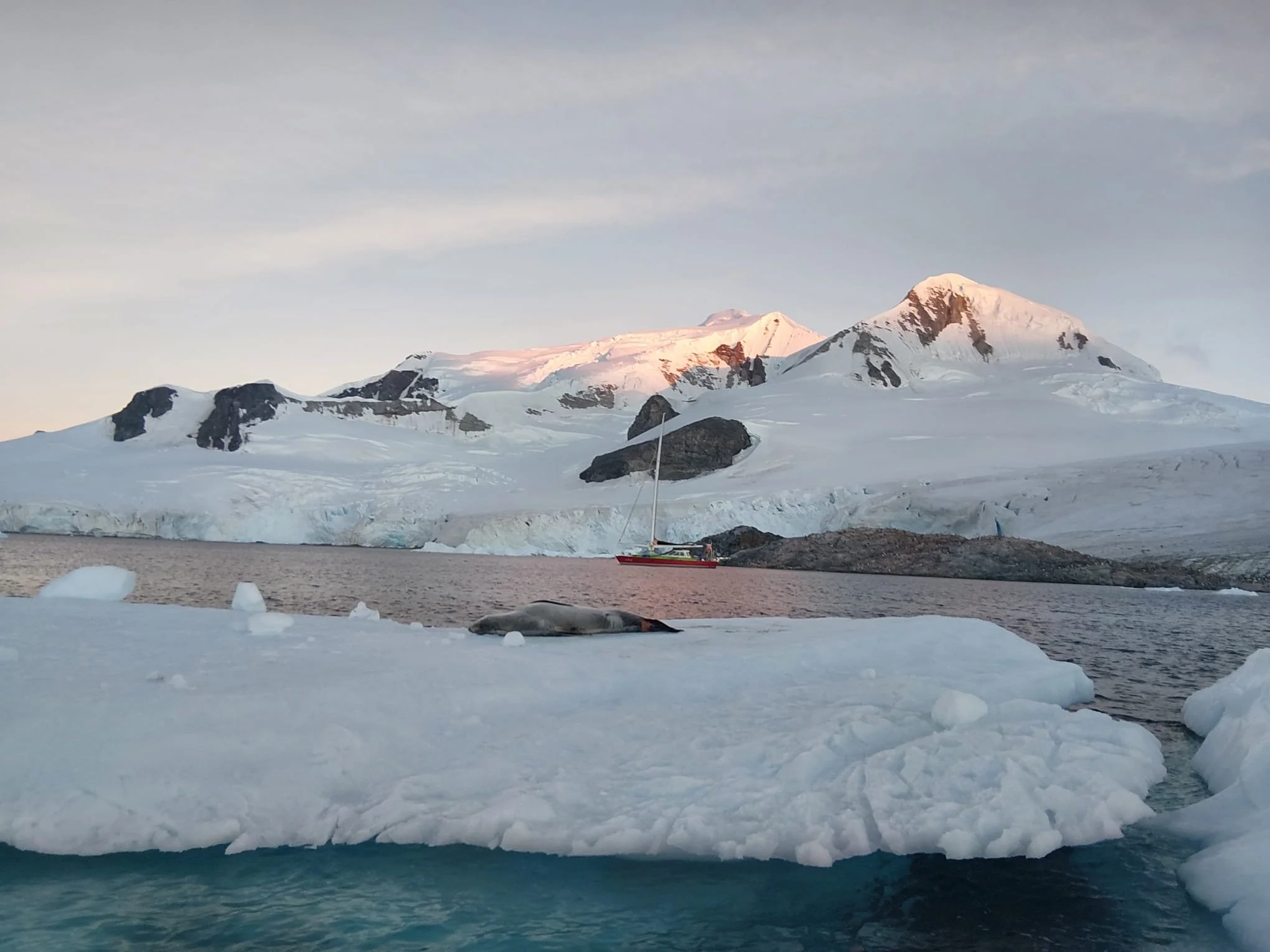 Snow-covered mountains with a glacier, a partially frozen body of water, and a sailboat near the shoreline during sunset.