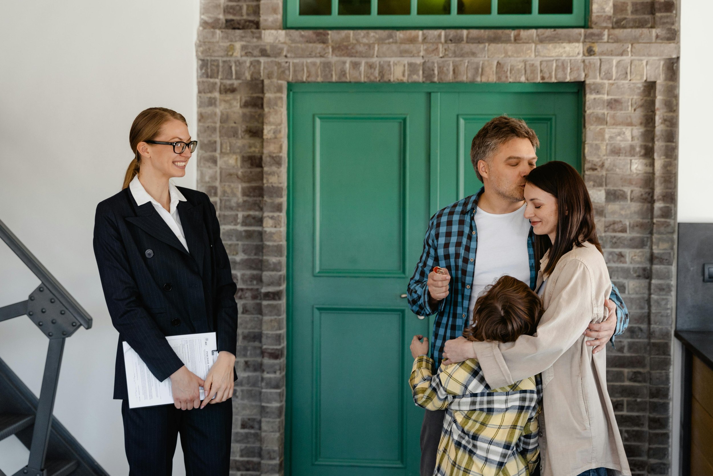 A family of three hugging with a woman and a young girl hugging while a man kisses the woman's forehead, and a woman in a black suit holding documents smiling nearby.