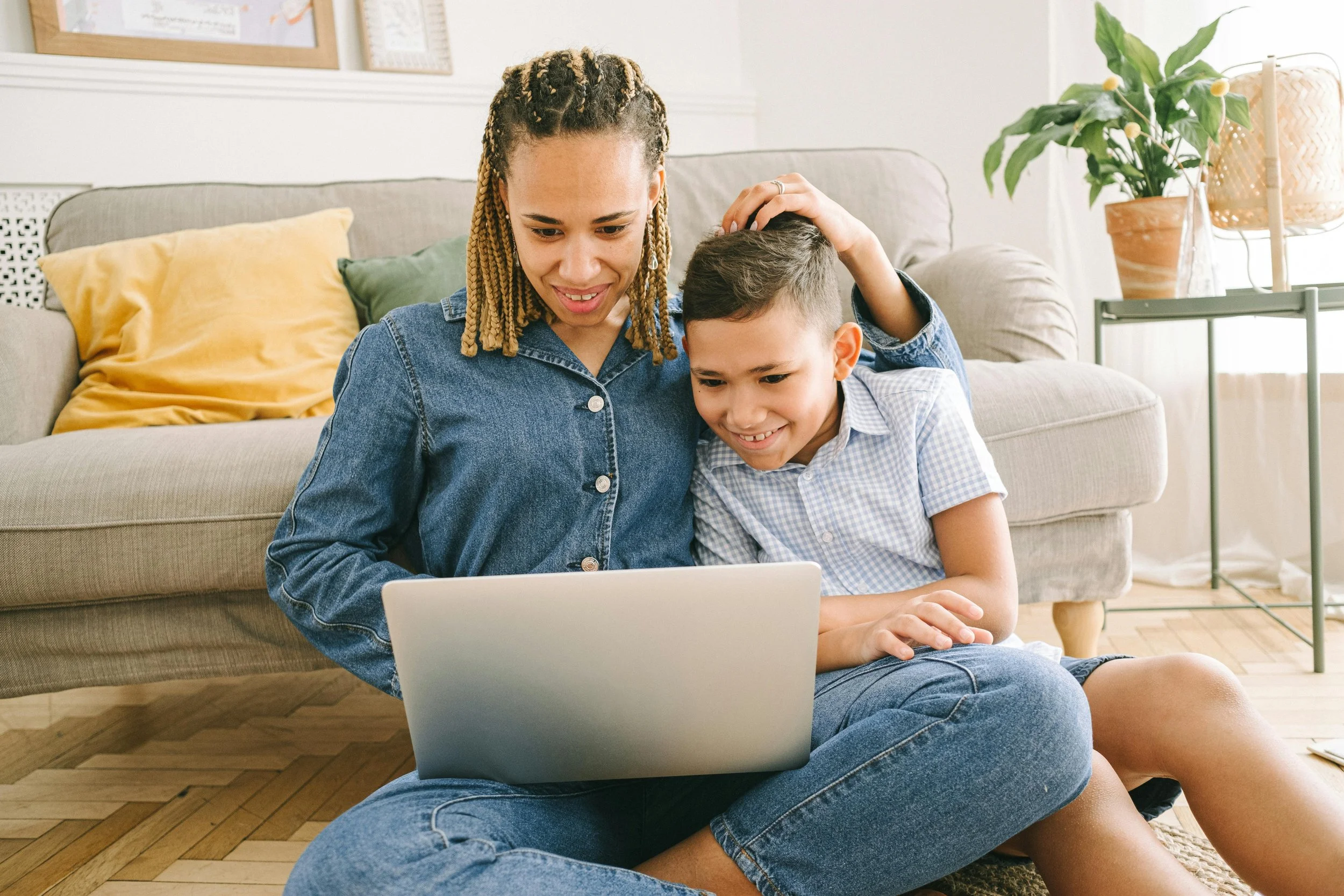 A woman and a young boy sitting on the floor in front of a laptop, smiling and looking at the screen together in a living room.