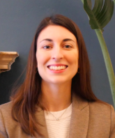 A woman with long brown hair smiling, wearing a beige blazer, with a large green plant and a blue wall in the background.