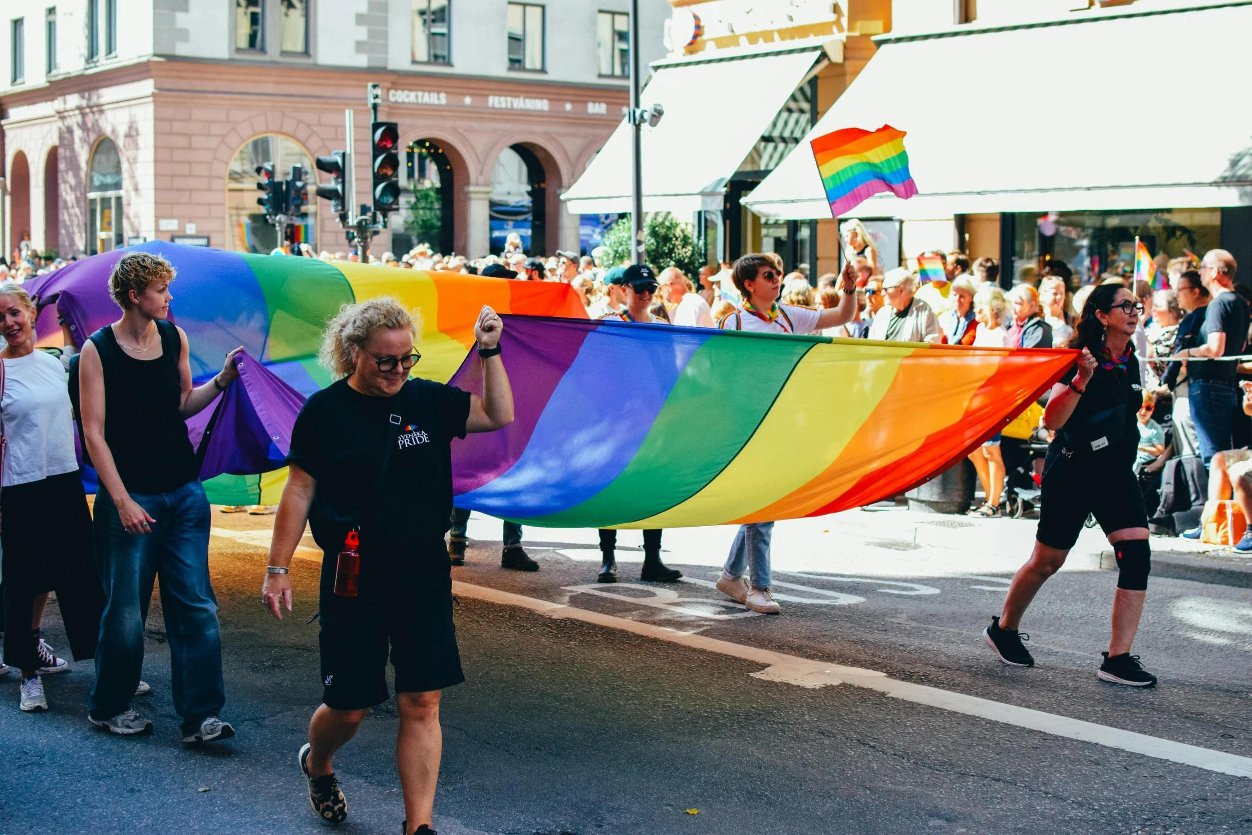 A group of people participating in a pride parade, holding a large rainbow flag. Many in the crowd are waving smaller rainbow flags, and some wear pride-themed accessories.