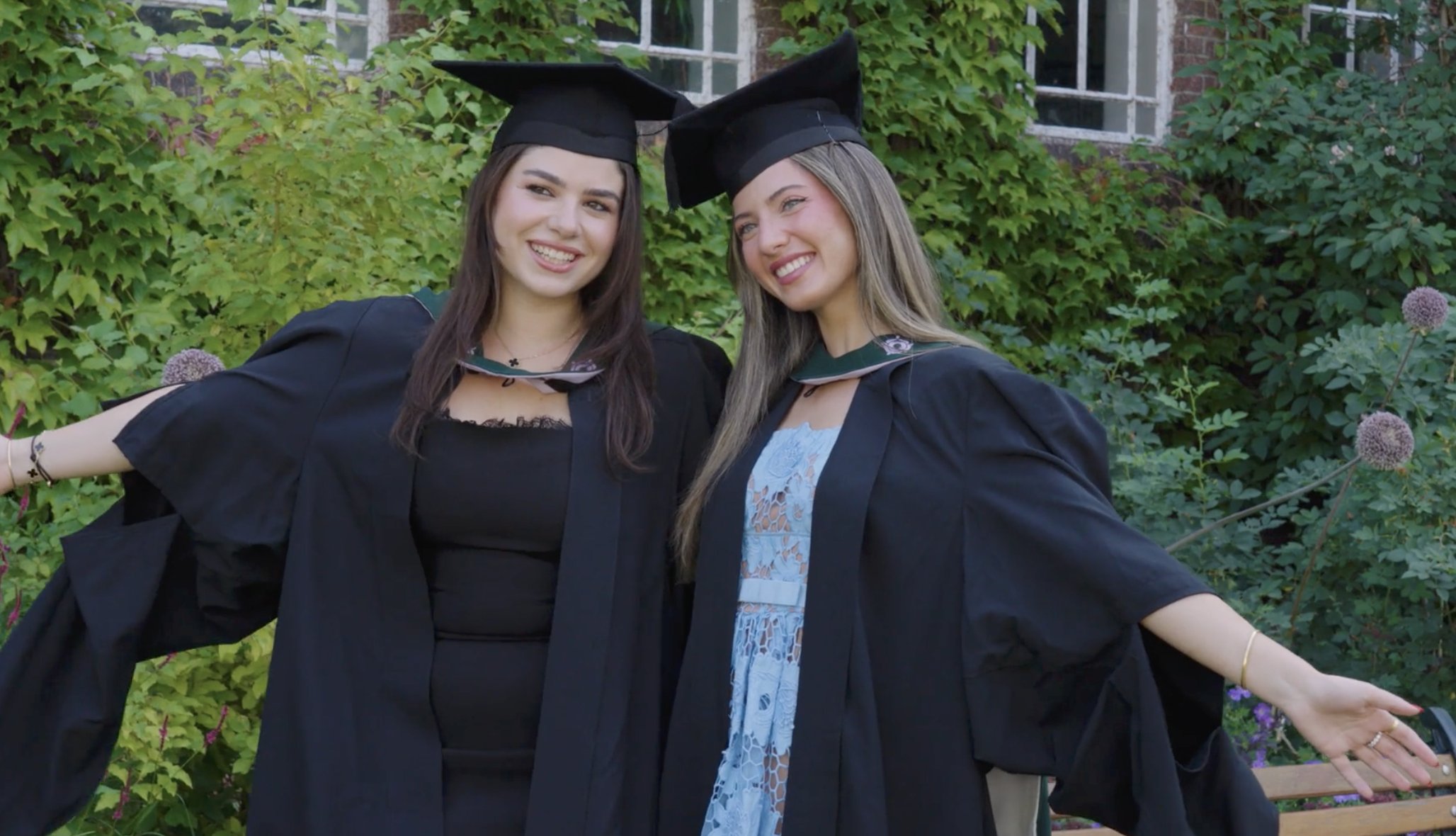 Two women dressed in black graduation caps and gowns smiling outdoors in front of a green leafy background.