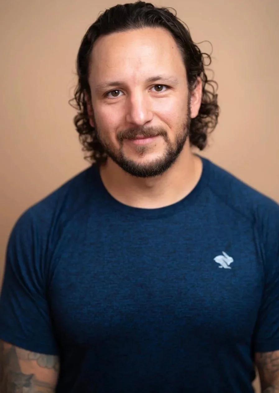 A man with shoulder-length curly hair, a beard, and a mustache, wearing a blue T-shirt with a small logo of a rabbit on the chest, smiling softly against a tan background.