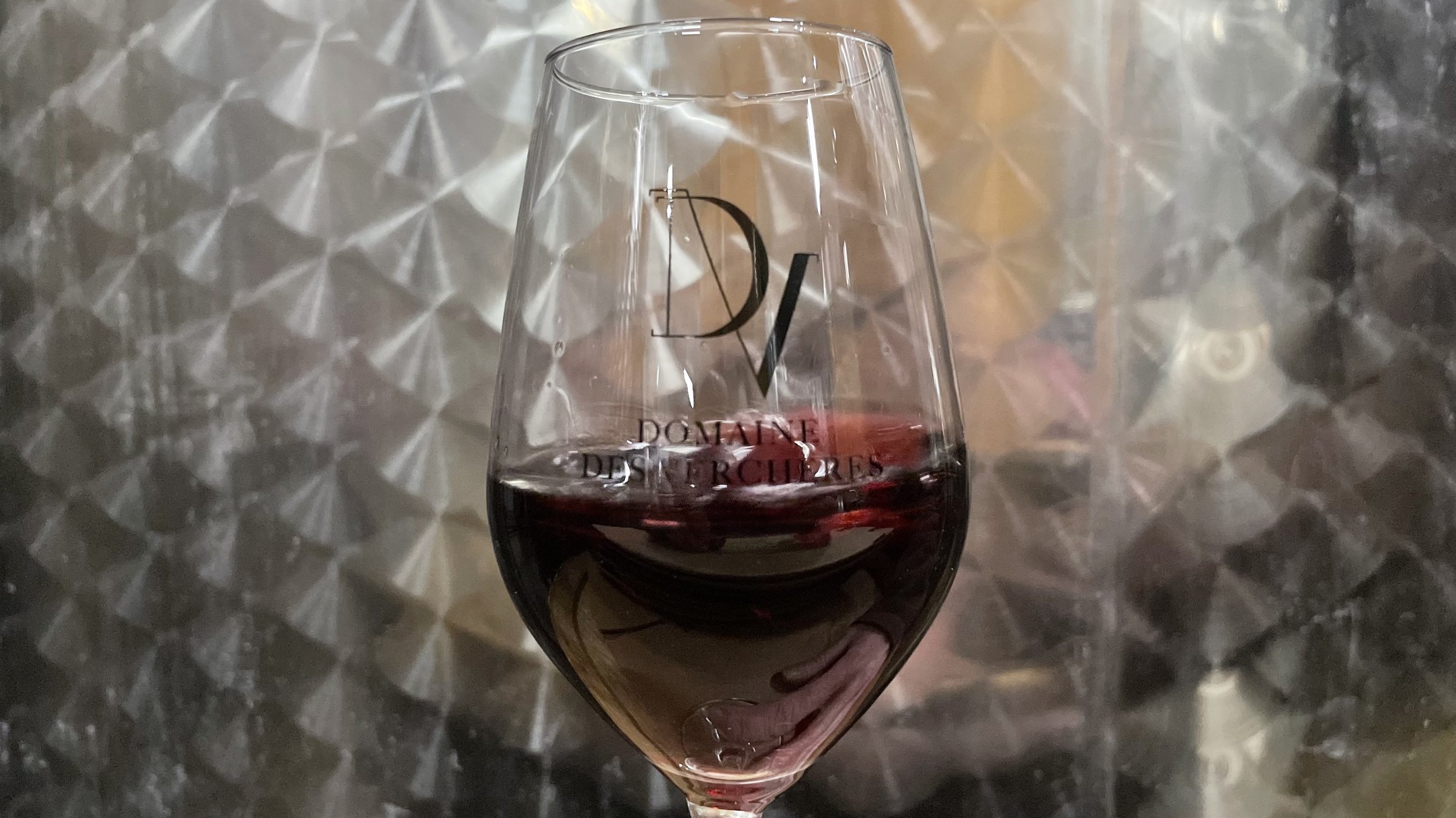Verre de vin rouge avec le logo 'Domaine des Verchères' en gros caractères noir, posé sur une surface métallique réfléchissante.