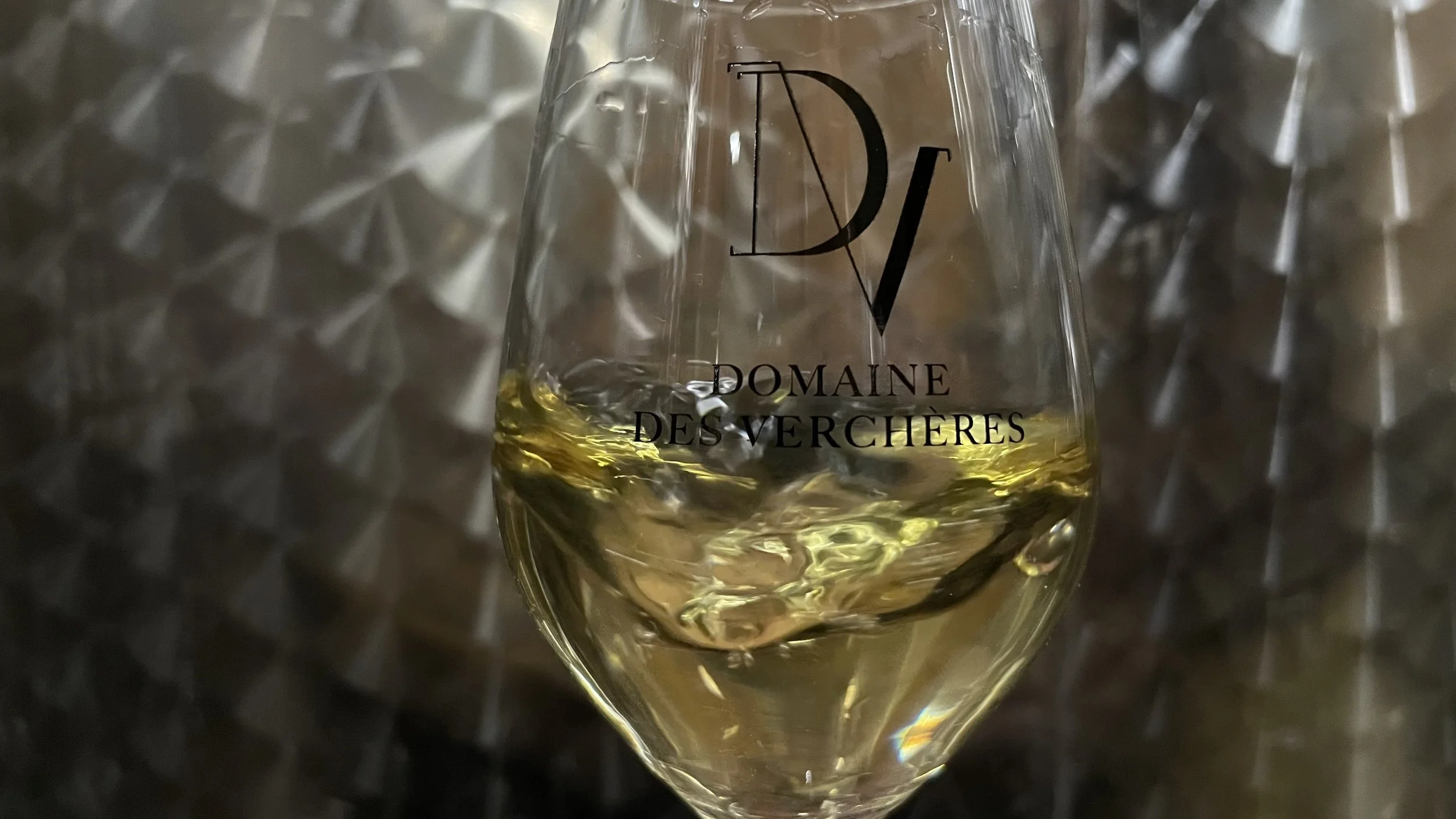 Verre de vin blanc avec le logo 'Domaine des Verchères' en gros caractères noir, posé sur une surface métallique réfléchissante.