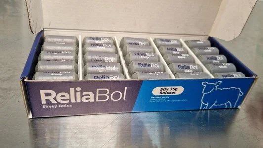 En åpen eske med flere små sylindriske reliekapsler, kalt ReliaBol Sheep Bolus, plassert på en metallbord.