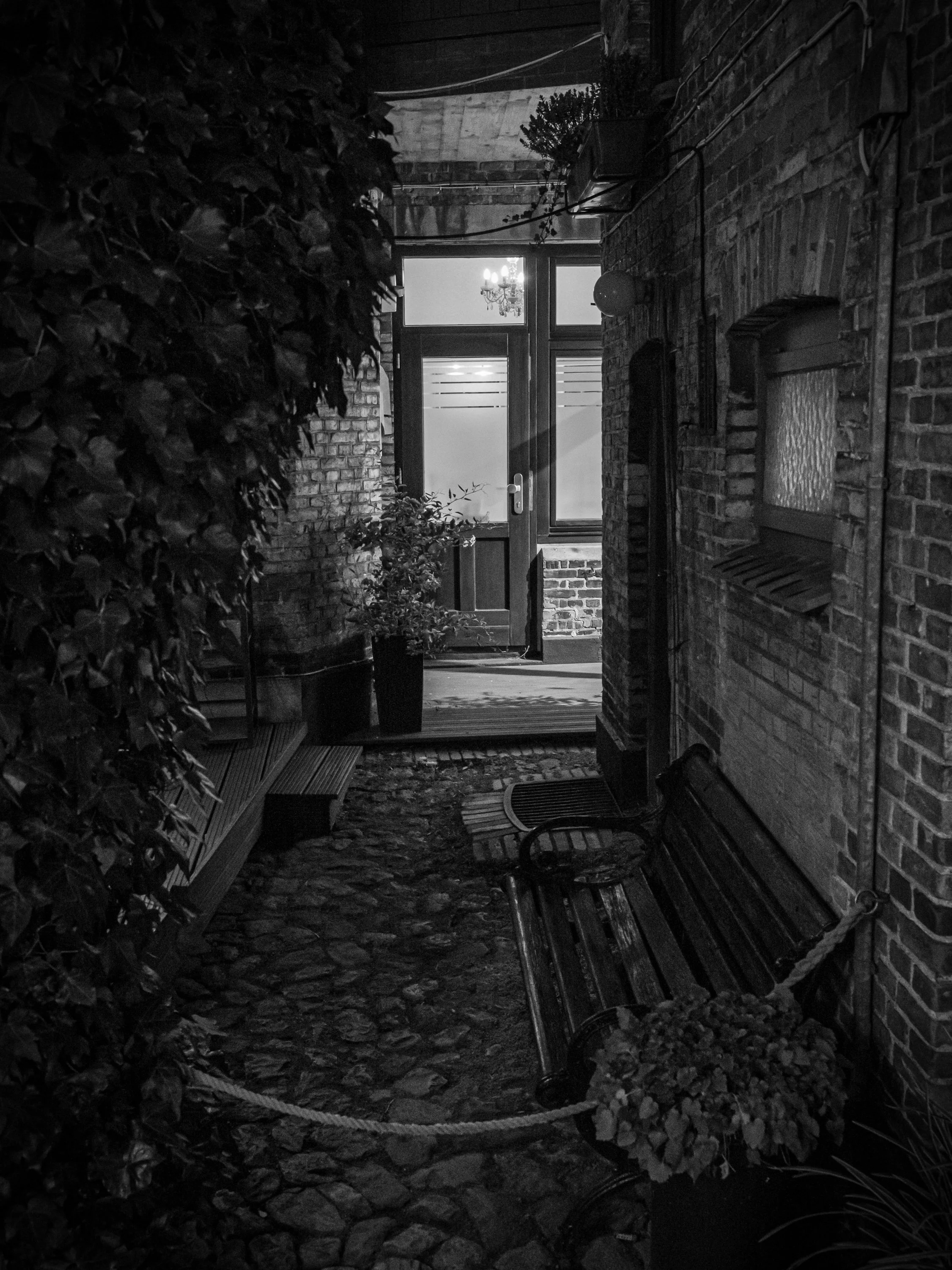 Narrow alley at night with brick walls, benches, and a lit doorway.