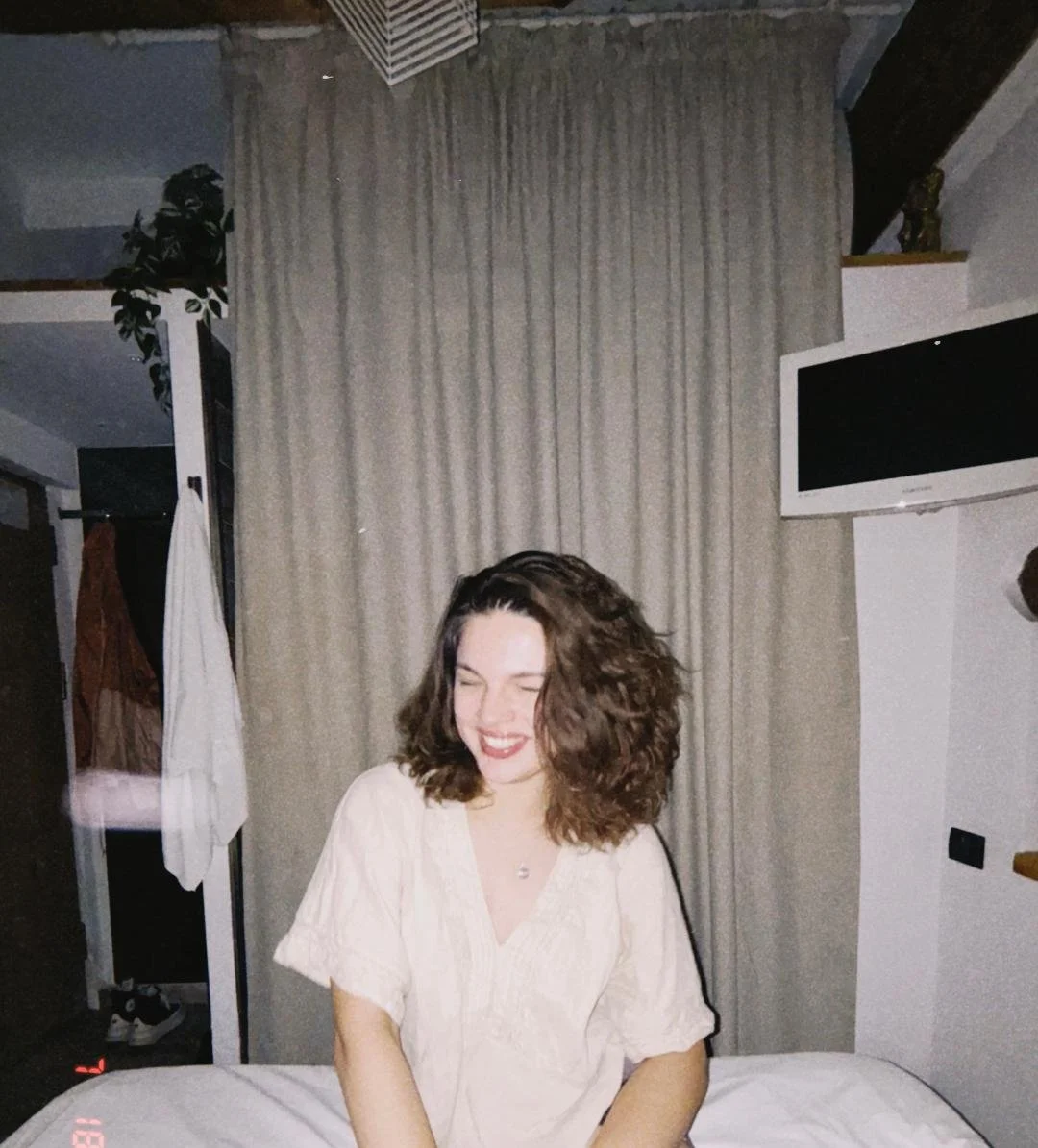 A young woman with curly brown hair sitting on a bed, smiling and laughing, in a room with beige curtains and a mounted TV.