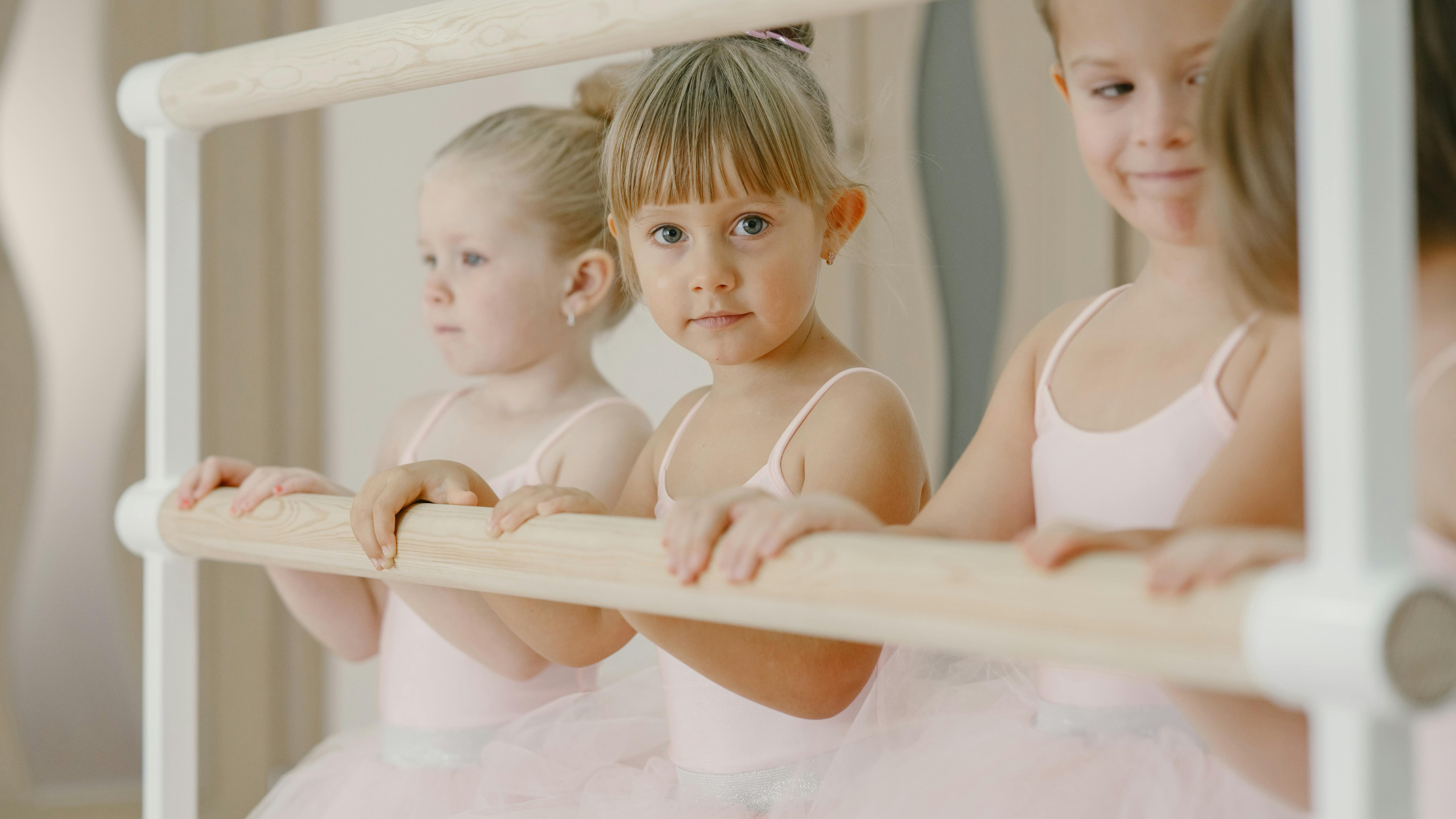 Three young girls in pink ballet costumes holding onto a ballet barre in a dance studio.