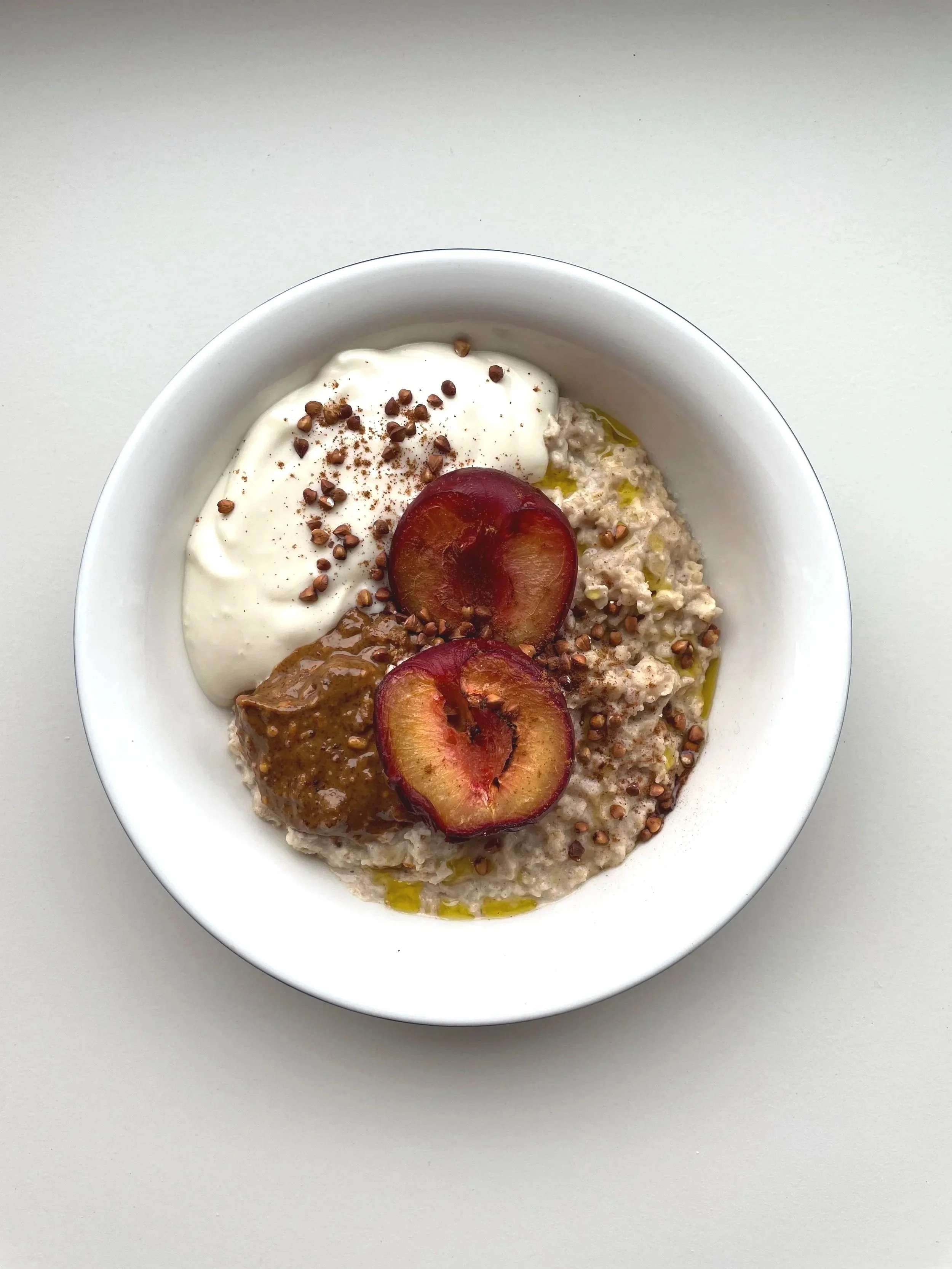A white bowl containing oatmeal topped with sliced plums, Greek yogurt, chia seeds, and a spoonful of peanut butter, drizzled with olive oil.