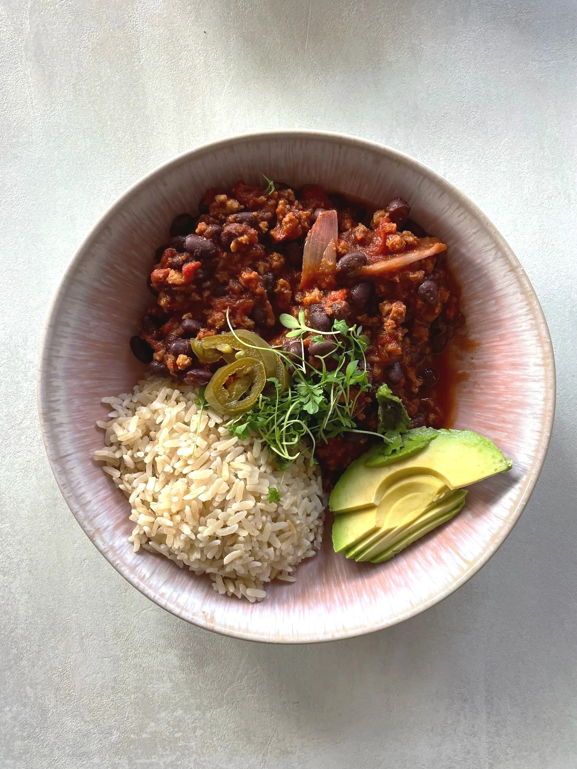 A bowl of rice, black bean chili with beef, sliced avocado, and garnished with microgreens and jalapenos.