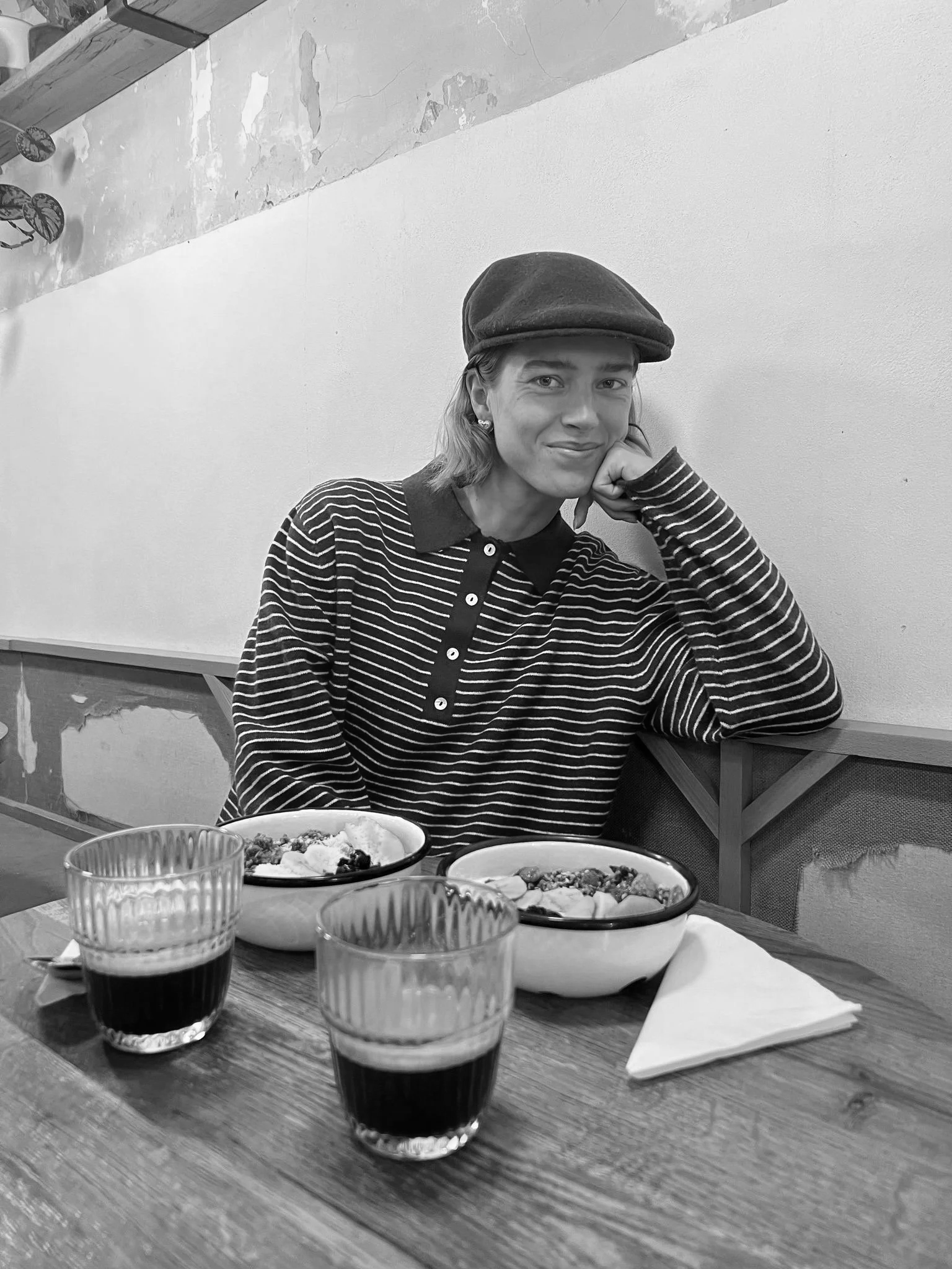 A woman sitting at a wooden table, resting her chin on her hand, with two bowls of dessert and two glasses of dark beverage in front of her, in a casual setting.