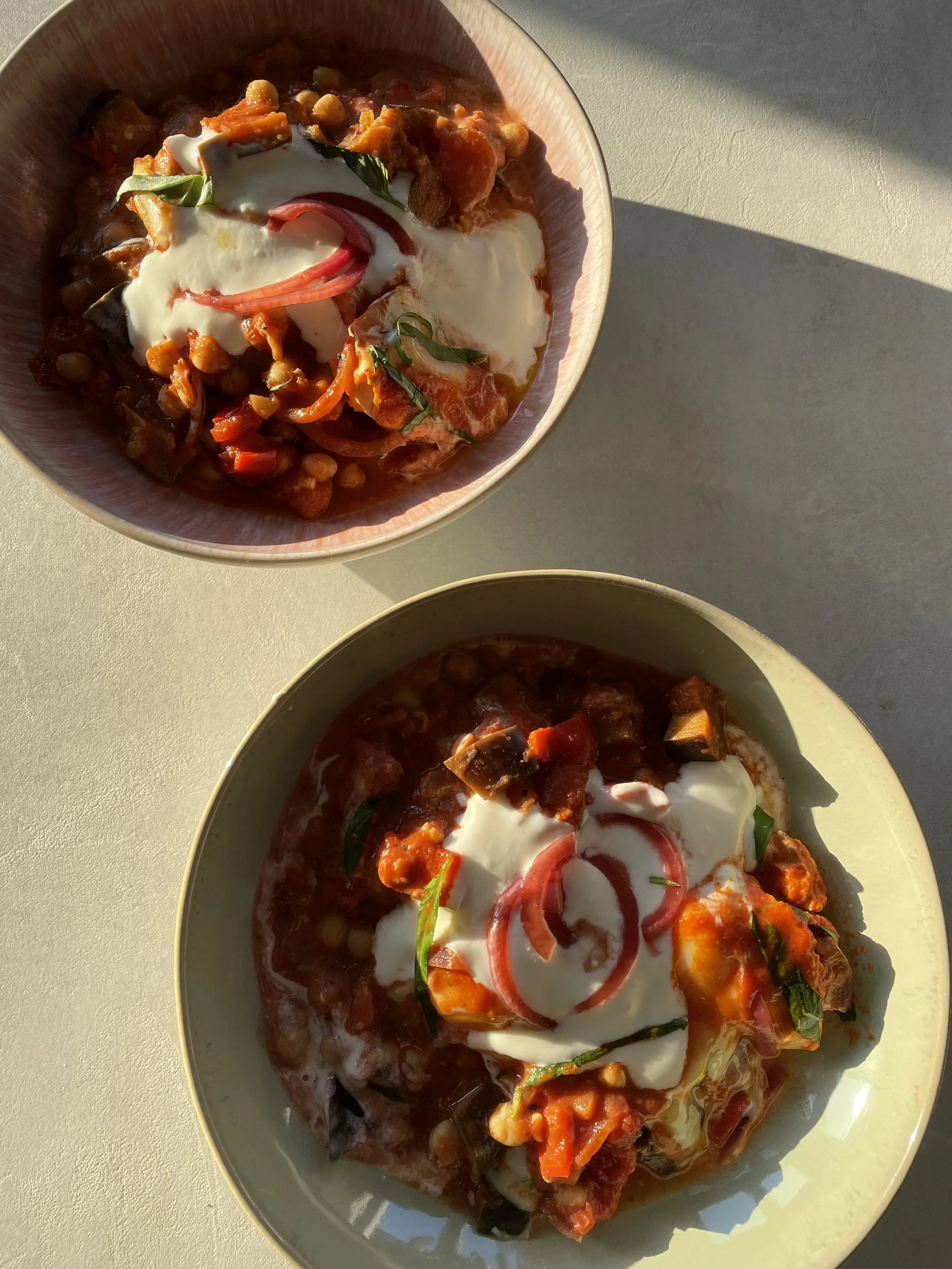Two bowls of vegetable stew topped with sour cream and sliced radishes, on a light-colored surface with sunlight shining on them.