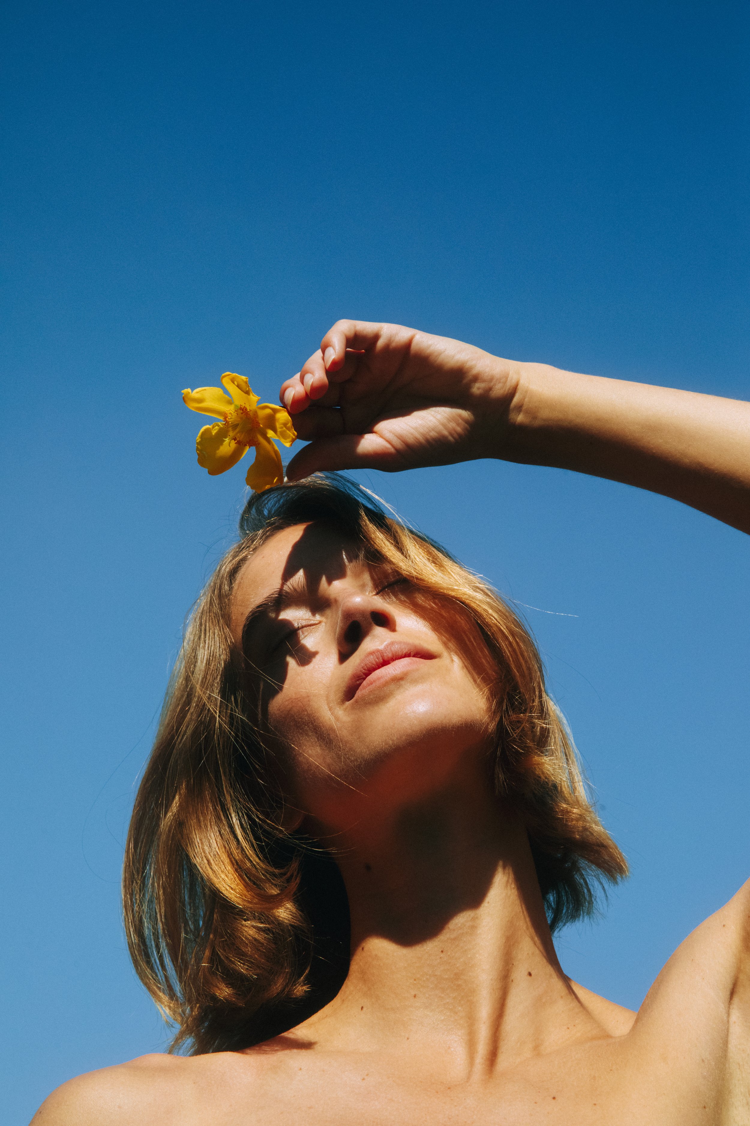 A woman with short brown hair holding a yellow flower above her head against a clear blue sky.