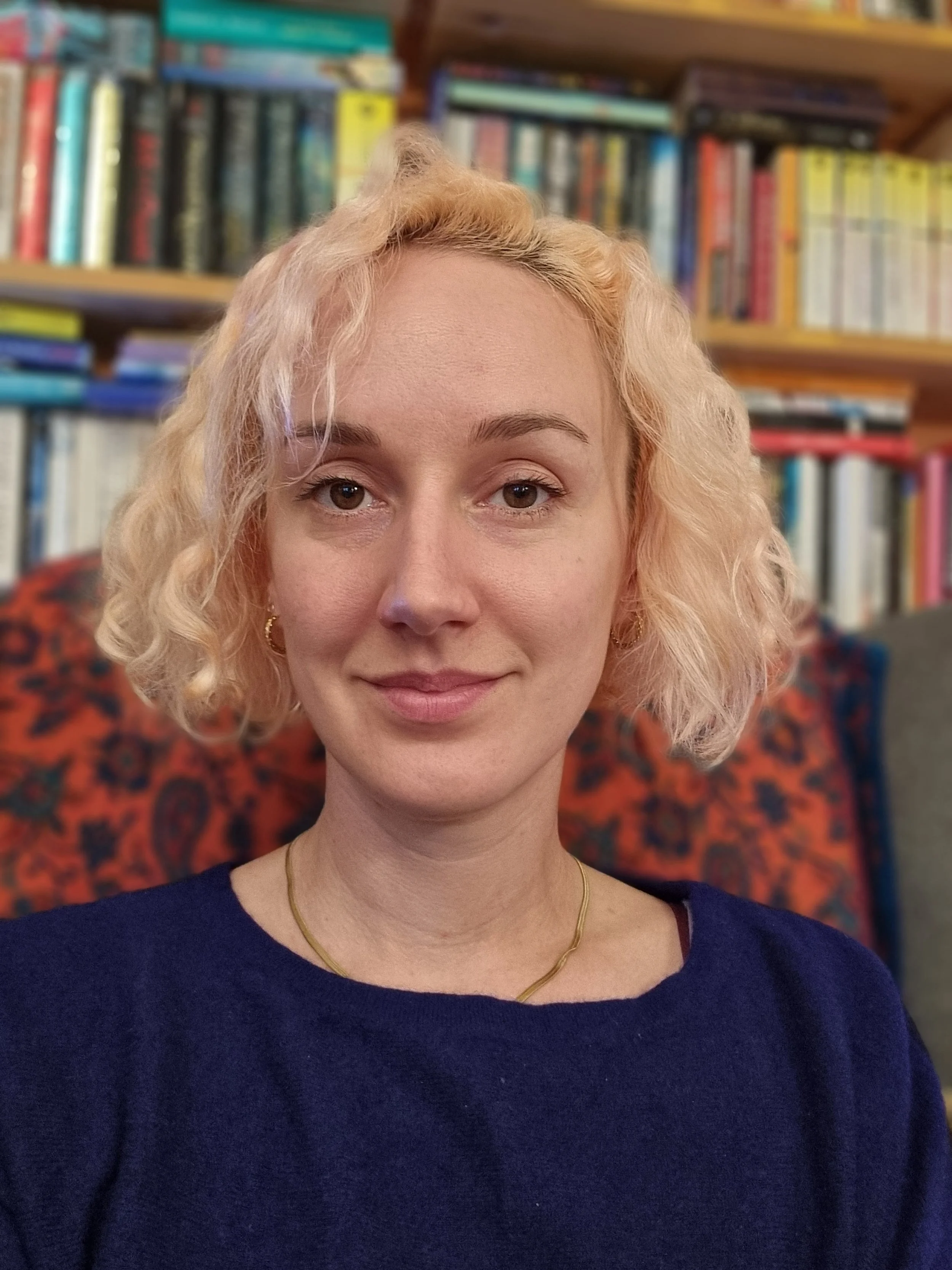 Psychosexual and relationship therapist Siân smiles at the camera - from the pink hair to the bookshelves in the background the picture is full of warmth and colour.