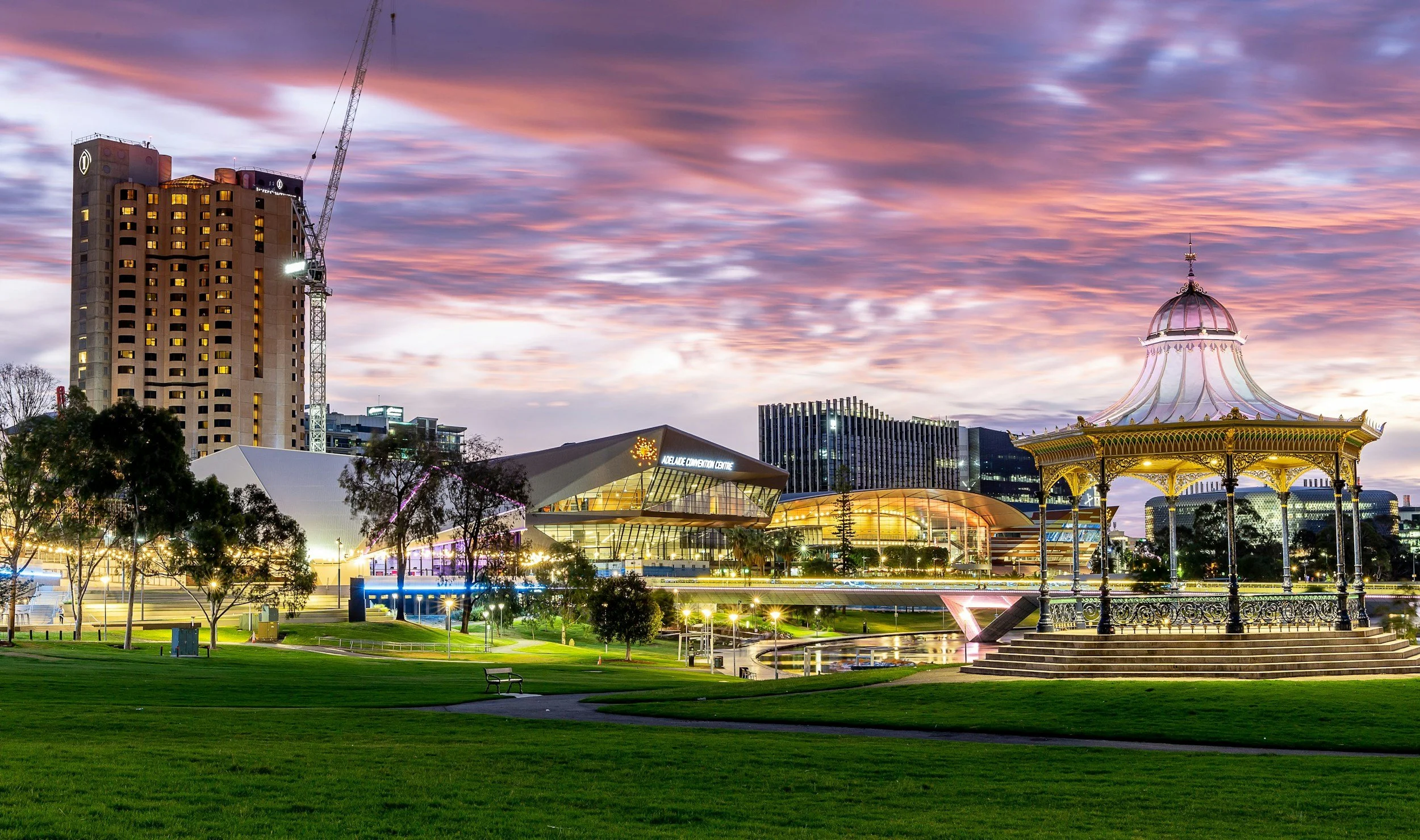 Night view of Perth cityscape with the Adelaide Entertainment Centre, a gazebo, and high-rise buildings under a colorful sunset sky.