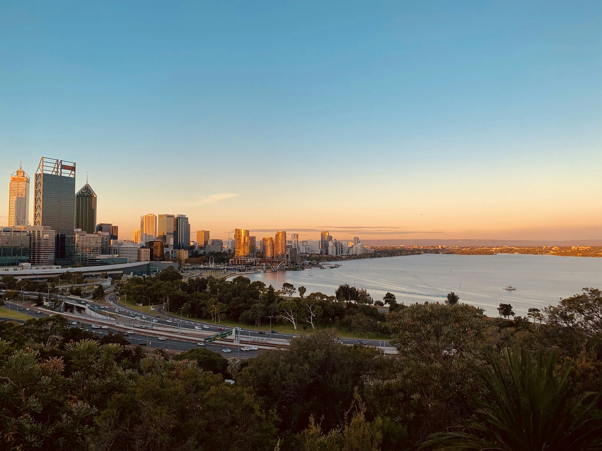 A city skyline at sunset with tall skyscrapers, a river, and lush greenery in the foreground.
