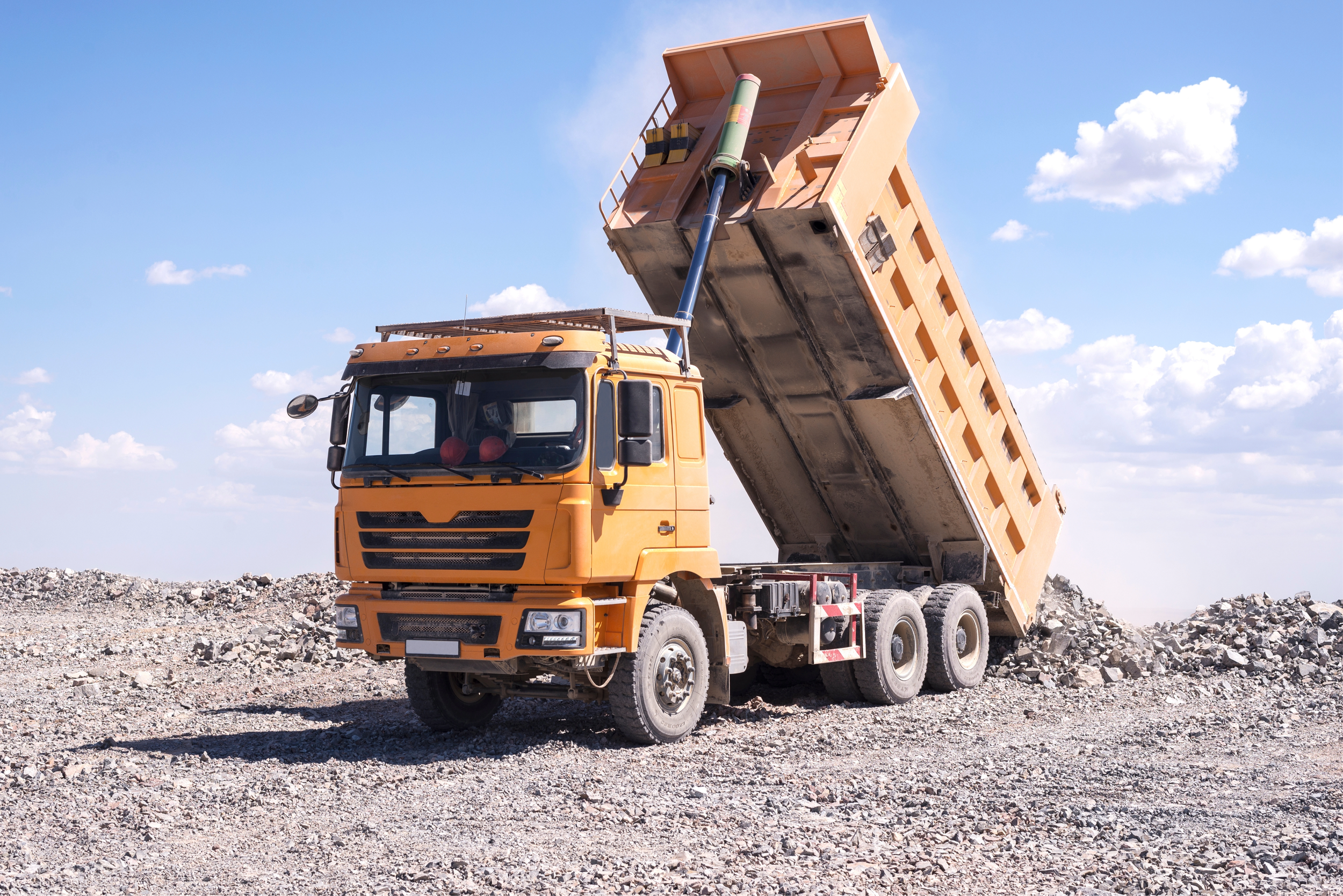 Dump truck with its bed raised, unloading a pile of gravel on a construction site under a blue sky with scattered clouds.