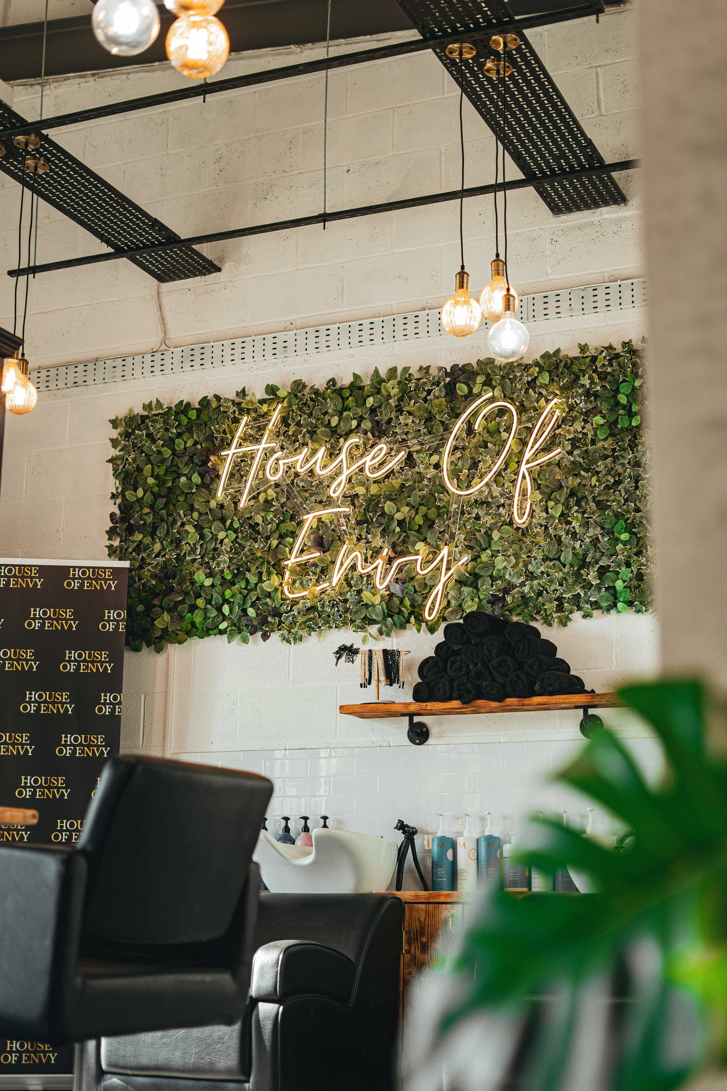 Interior of a modern hair salon with a neon sign that reads 'House Of Envy' on a green leafy wall, black salon chairs, hair washing stations, and hanging light bulbs.