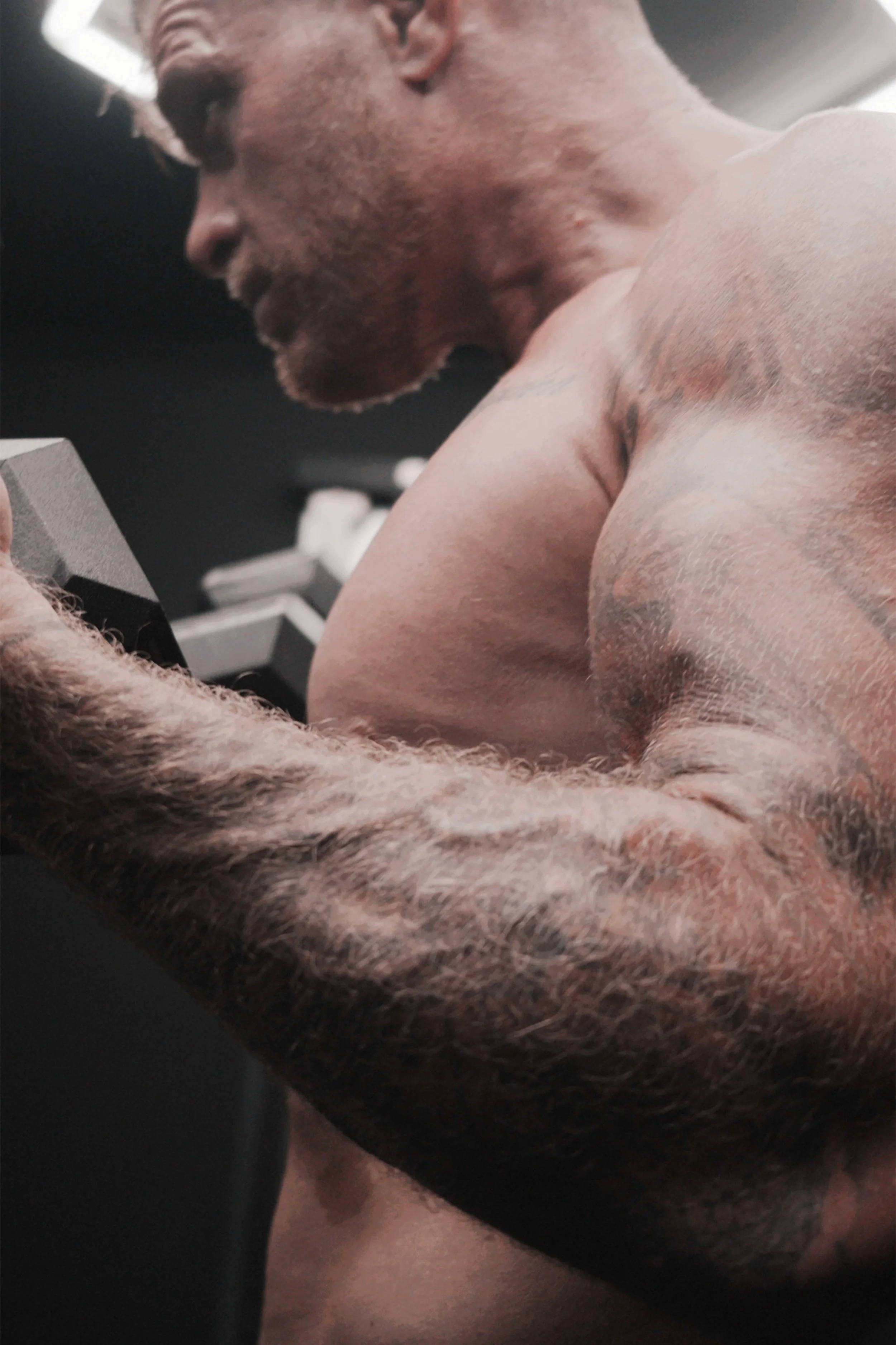A muscular man with tattoos on his arm and chest, shirtless, looking down while in a gym or workout setting.