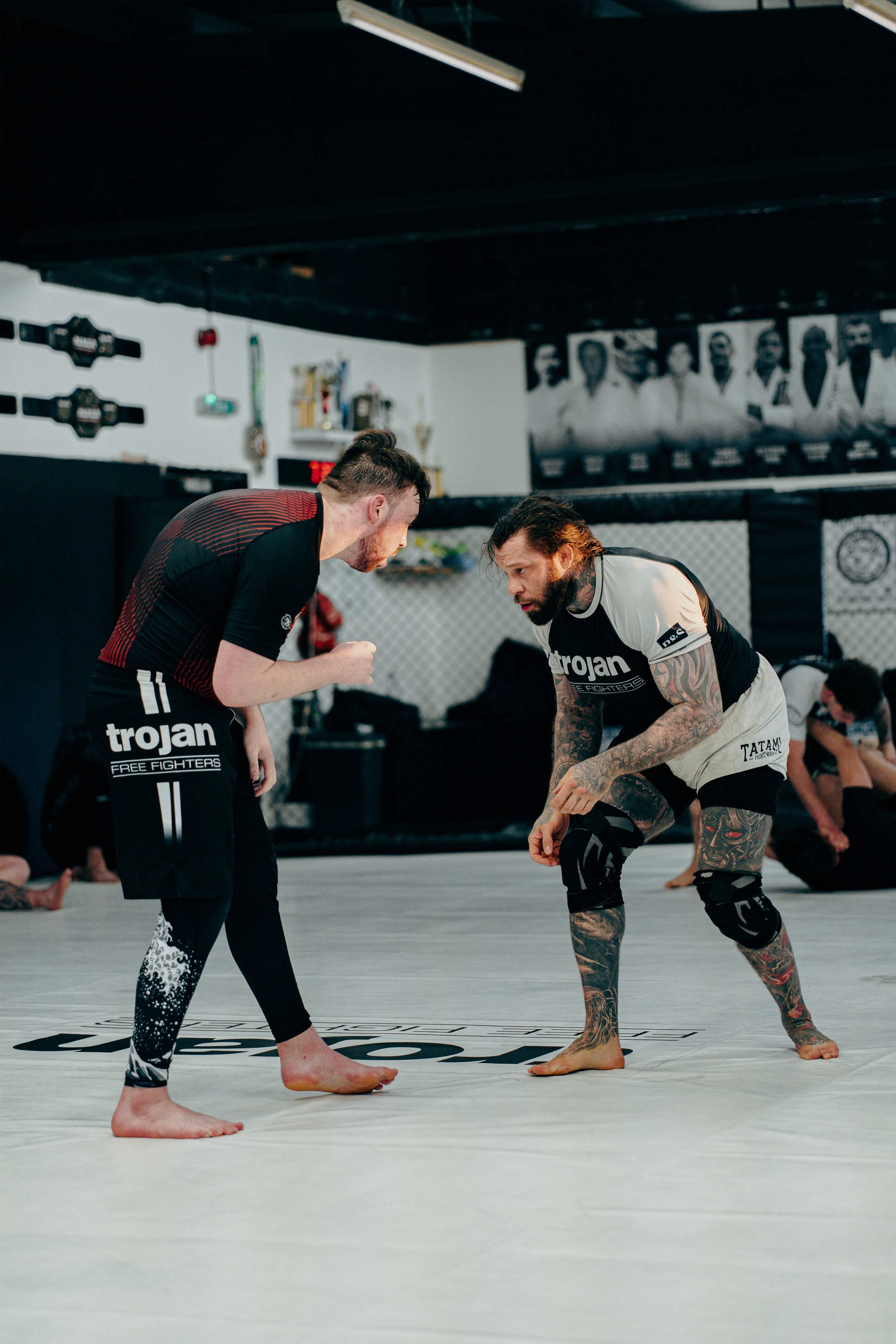 Two male fighters training in a gym inside an MMA training facility, one in black with red stripes and the other with tattoos, wearing rash guards and shorts, engaged in a grappling drill on the mat.