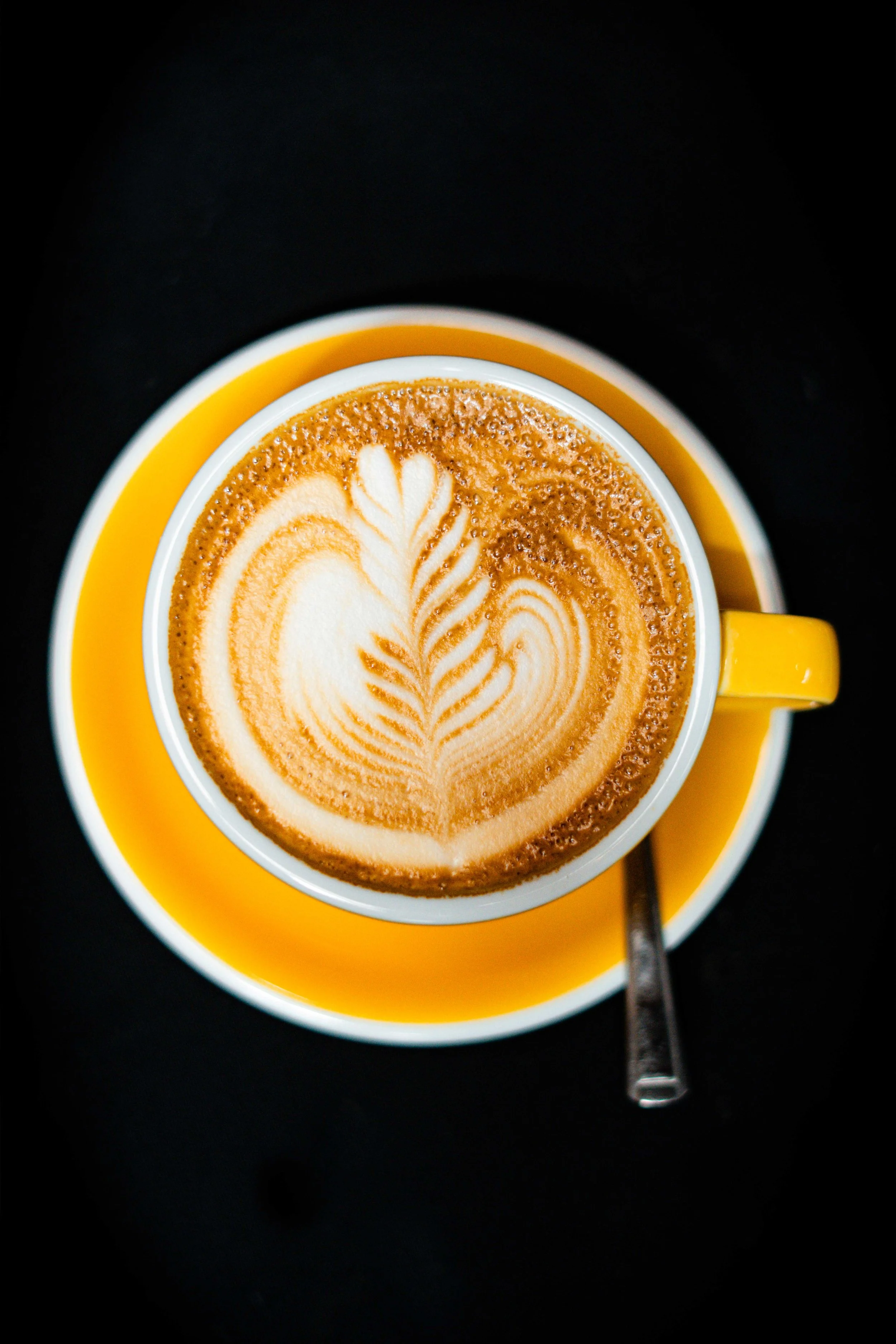 A cup of coffee with latte art on top, placed on a yellow saucer with a metal spoon on a black surface.