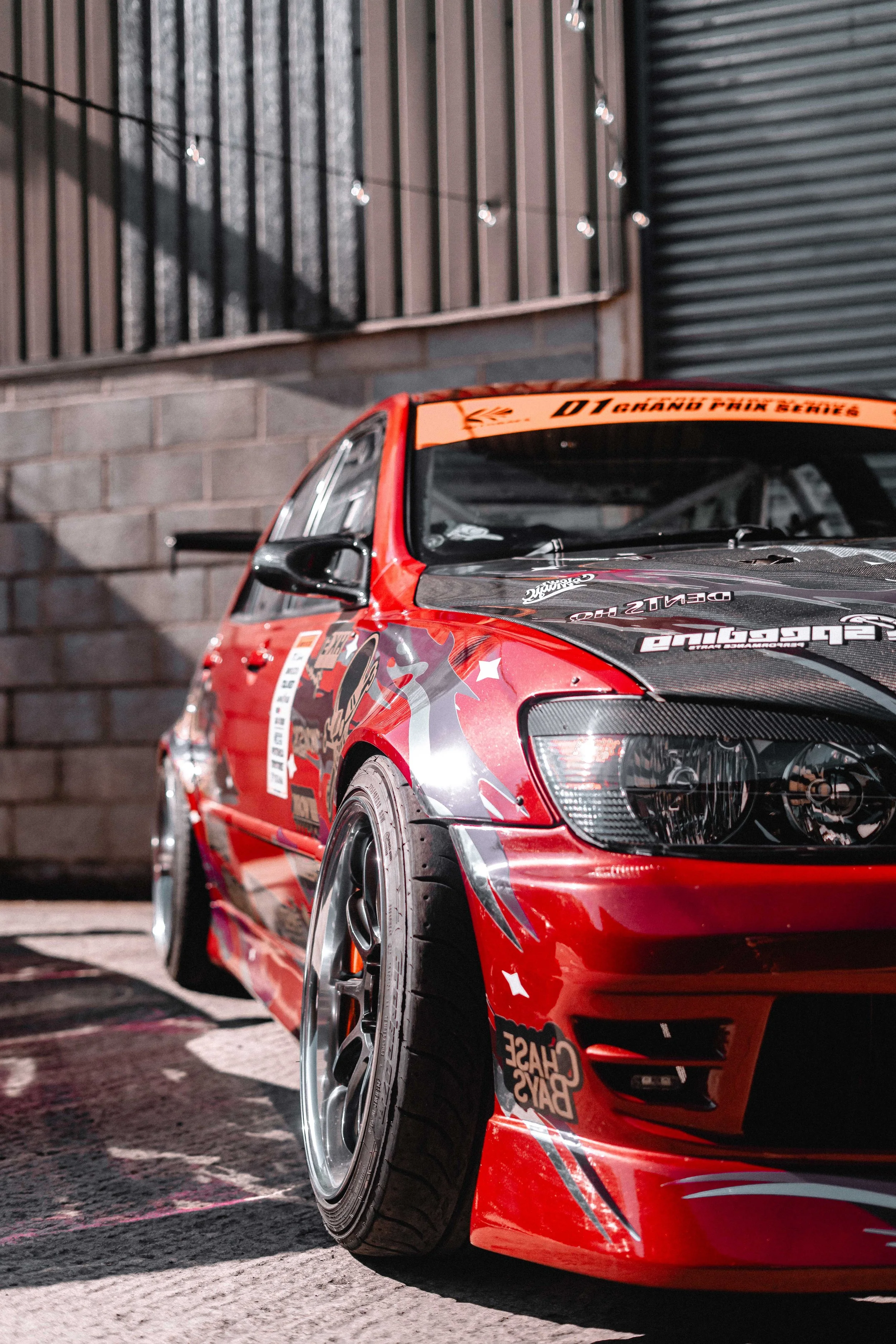 A red race car with black and white graphics, parked outside near a brick wall and a metal door, featuring a front bumper and custom wheels.