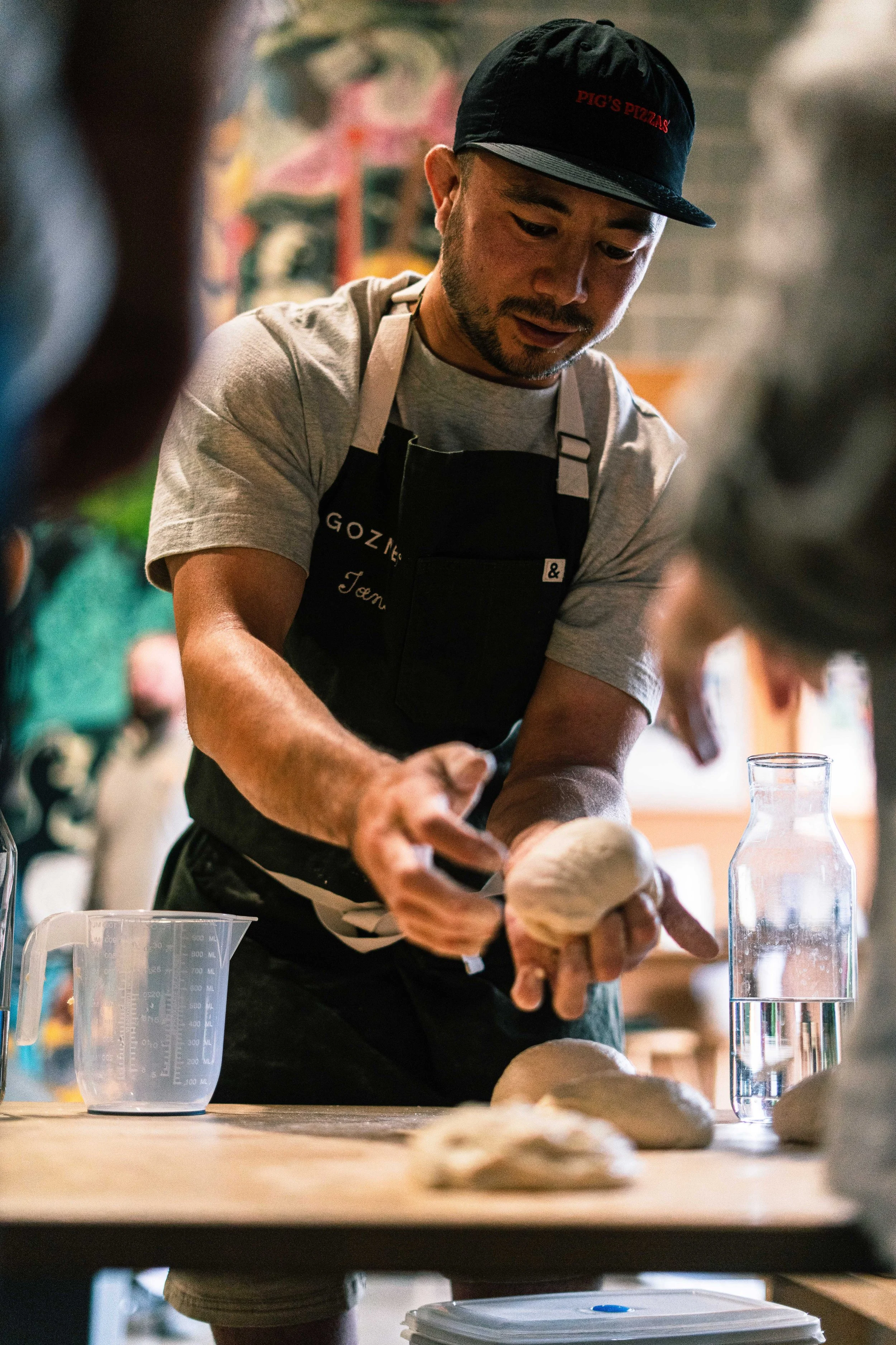 A man wearing a black cap and apron is shaping dough on a wooden table in a bakery or kitchen.