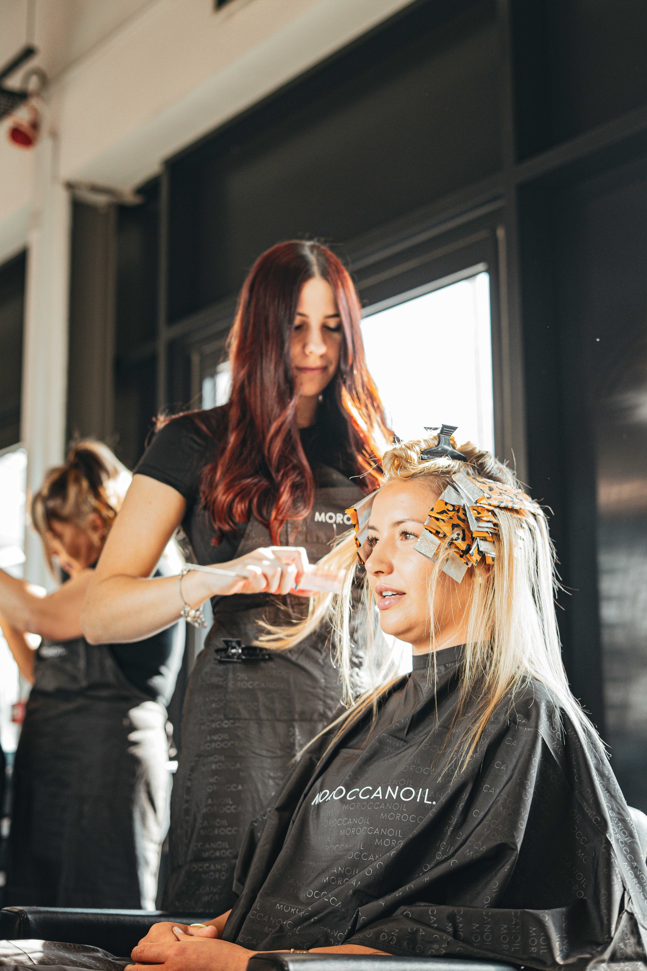 A hairstylist applying hair dye to a woman's hair who is sitting in a salon chair with rollers and foil in her hair, and another hairstylist working in the background.