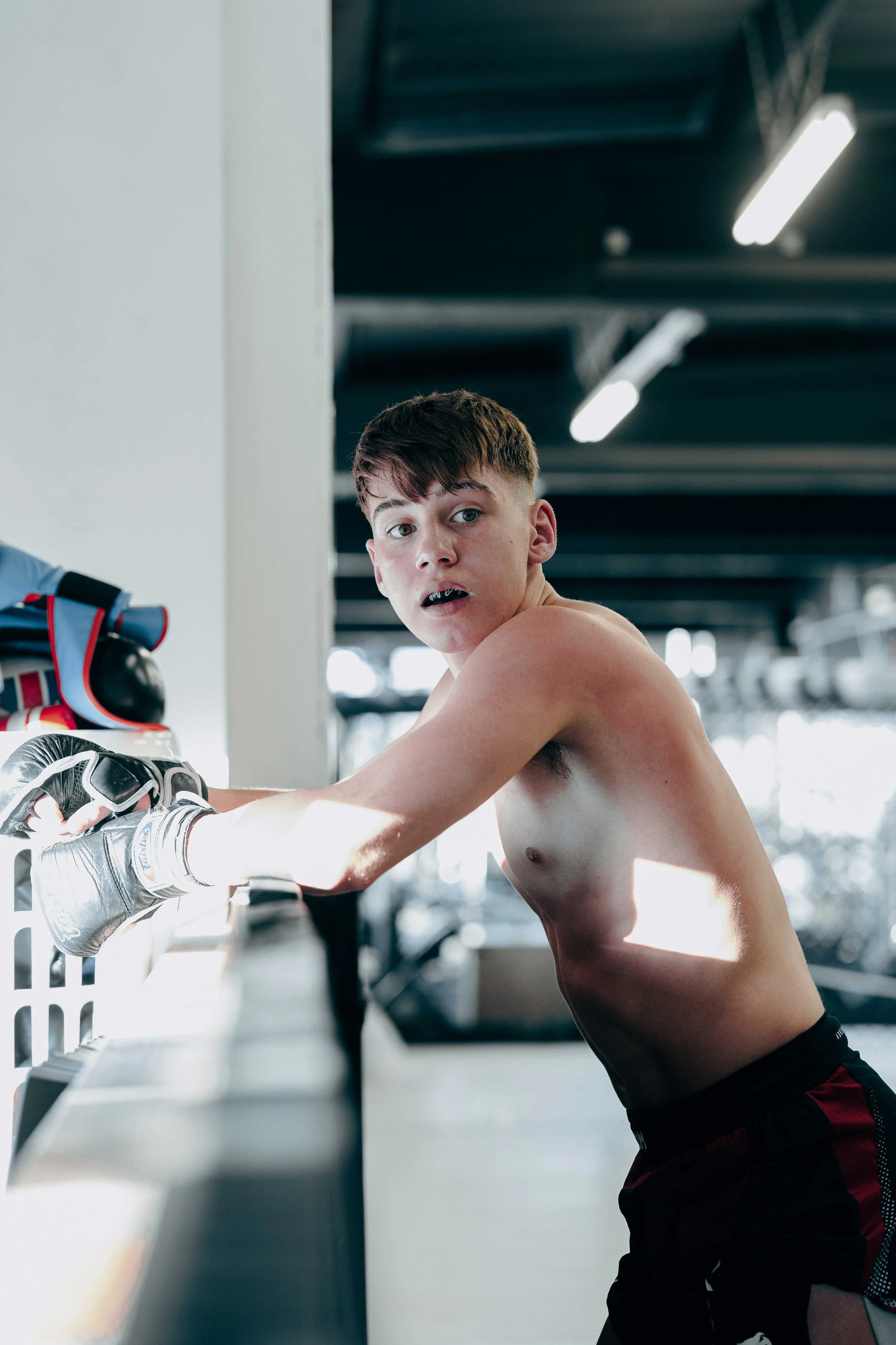A shirtless young man with short hair and a surprised expression, wearing black gloves, leaning on a counter in a gym or training facility.