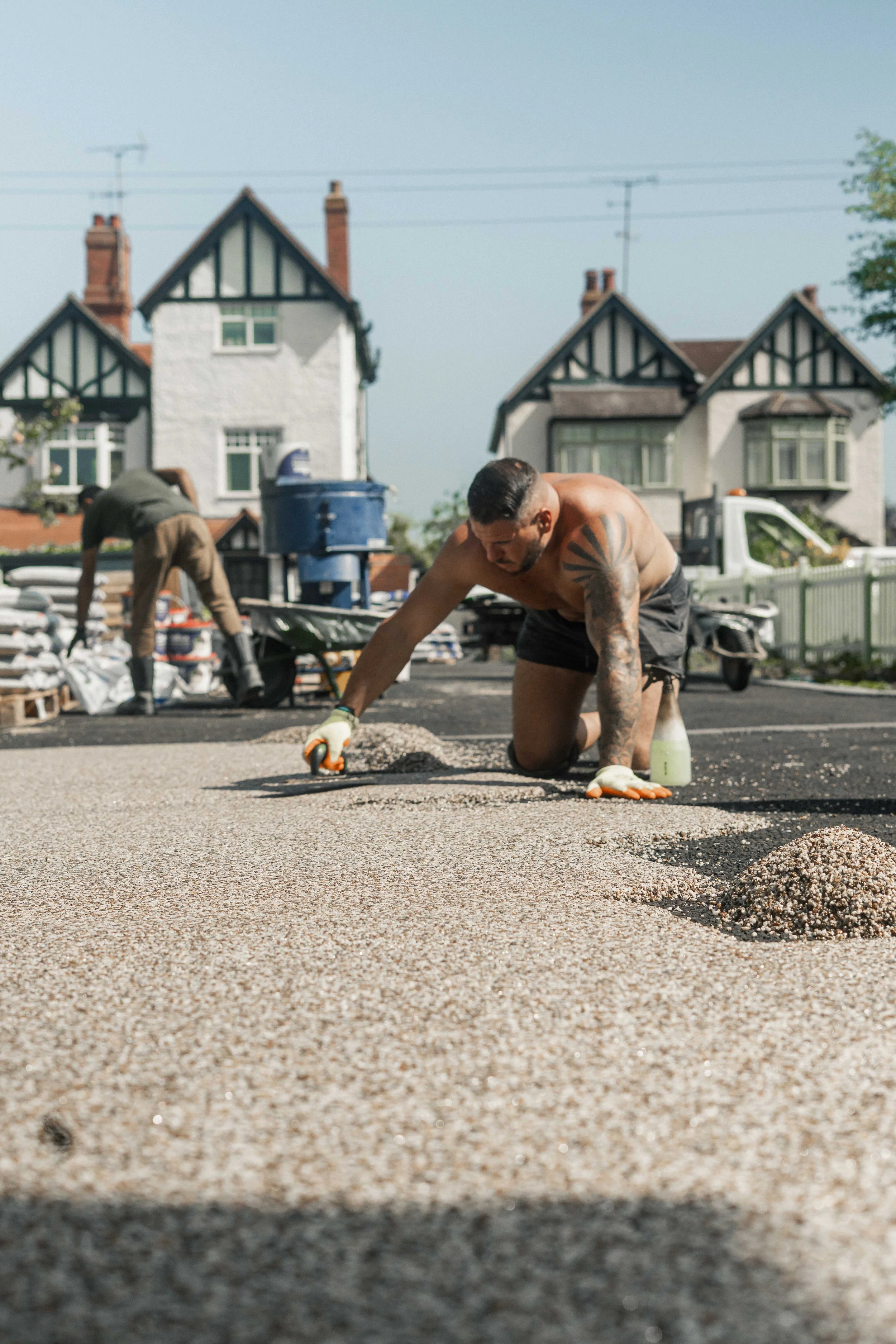 A man working on a construction project laying gravel or sand on a road surface. He is kneeling on the ground, wearing gloves, black shorts, and has tattoos on his arm. In the background, another person is working, with houses and a white picket fence visible.