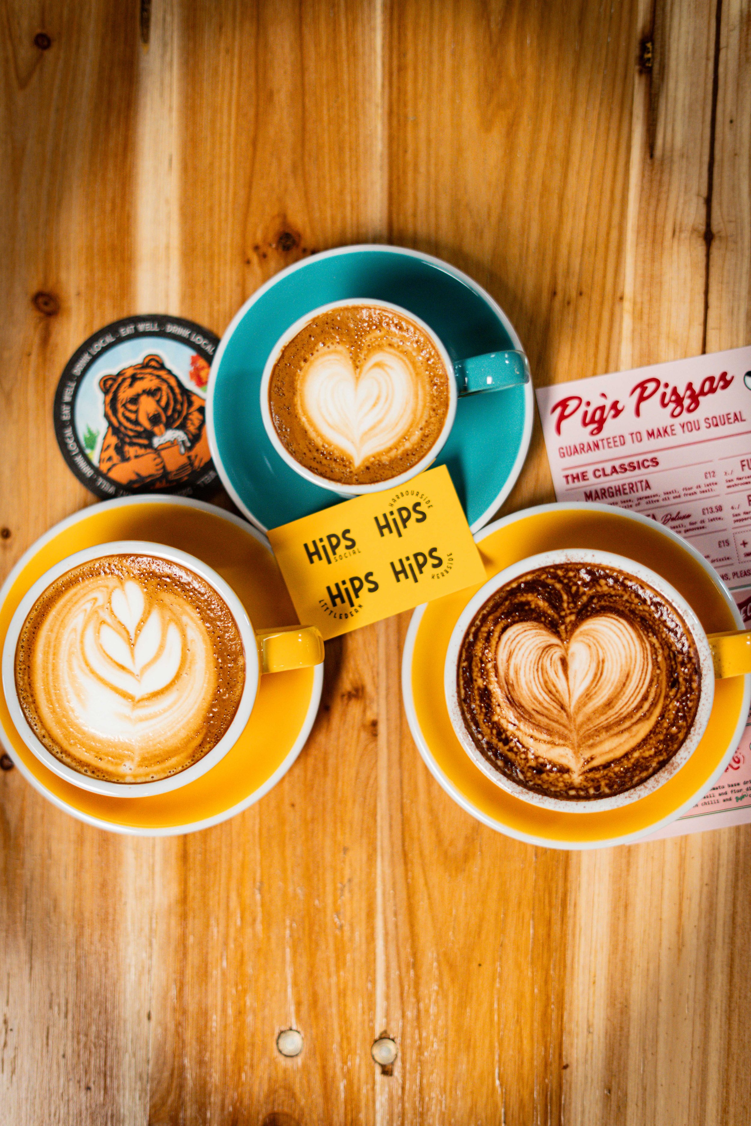 Three cups of coffee with latte art, set on a wooden table, with a pig pizza menu and yellow HipS sticky notes in the center.