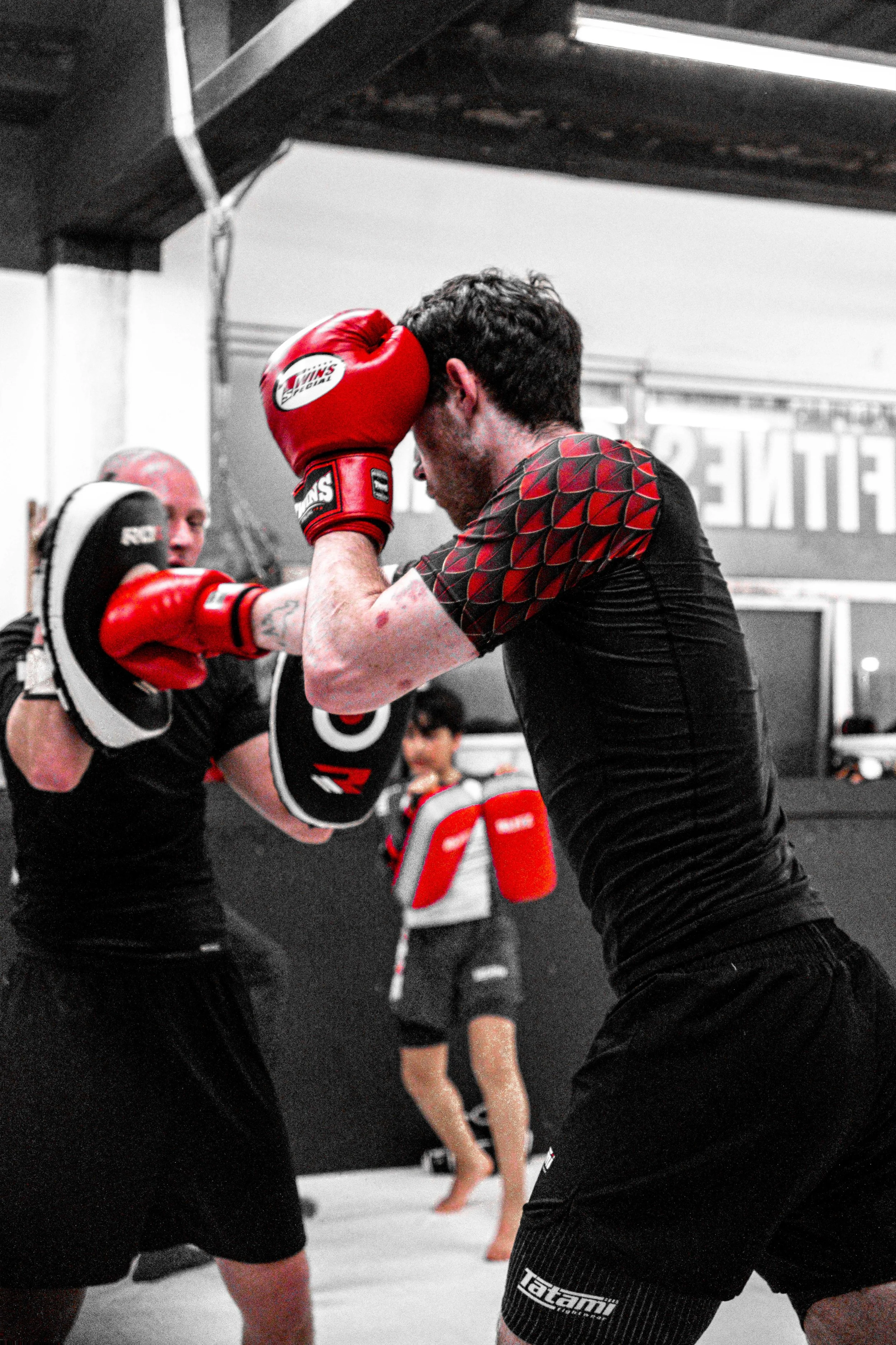 Two men practicing boxing in a gym, wearing red boxing gloves and black athletic clothing, with a young boy in the background holding boxing mitts.