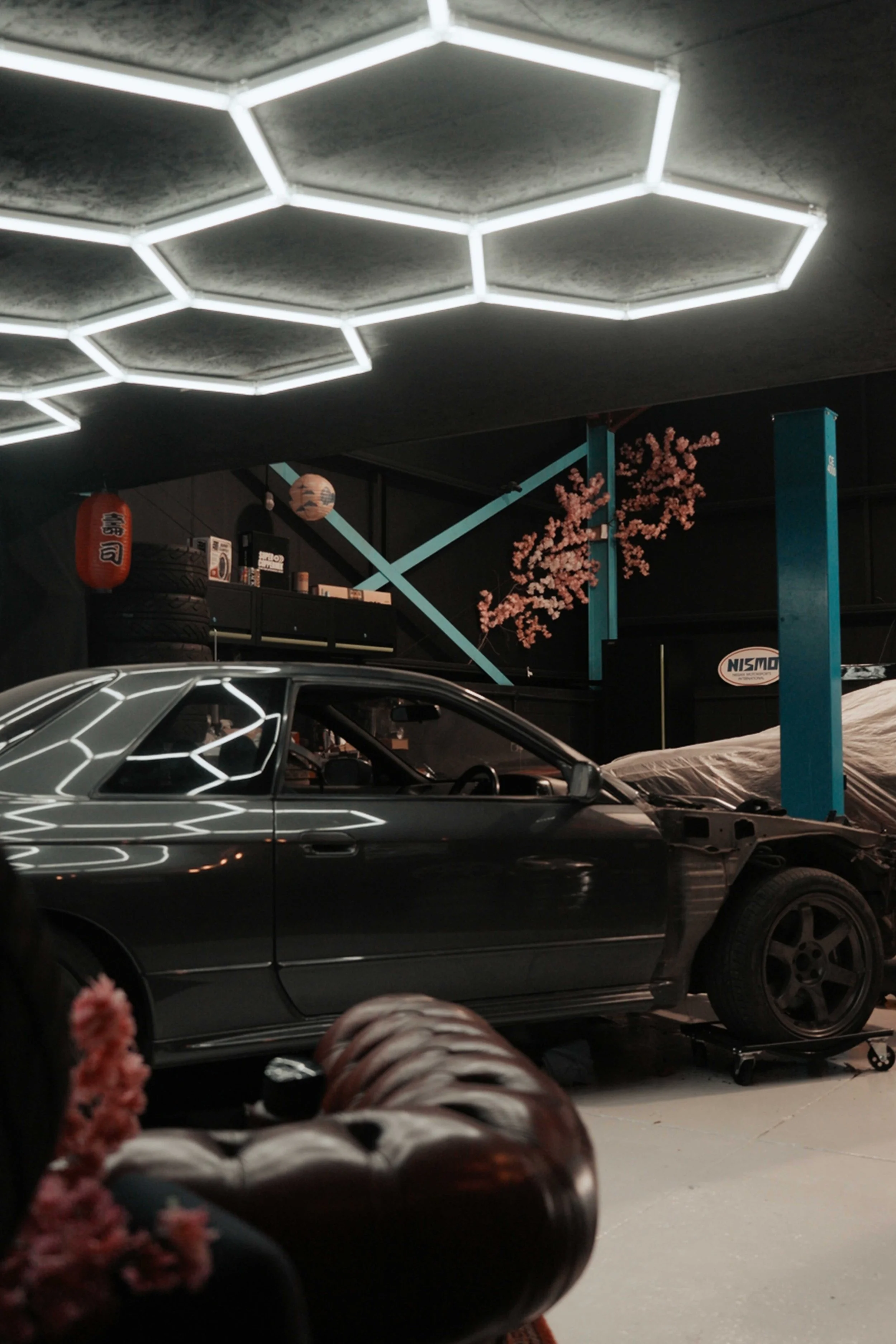 An indoor space with a modern hexagon-shaped LED ceiling light. There is a black car with some parts missing, like the front bumper, on a lift or jack. The background has Japanese paper lanterns, tires, and shelves with small items. Pink cherry blossom decorations are also visible on the wall.