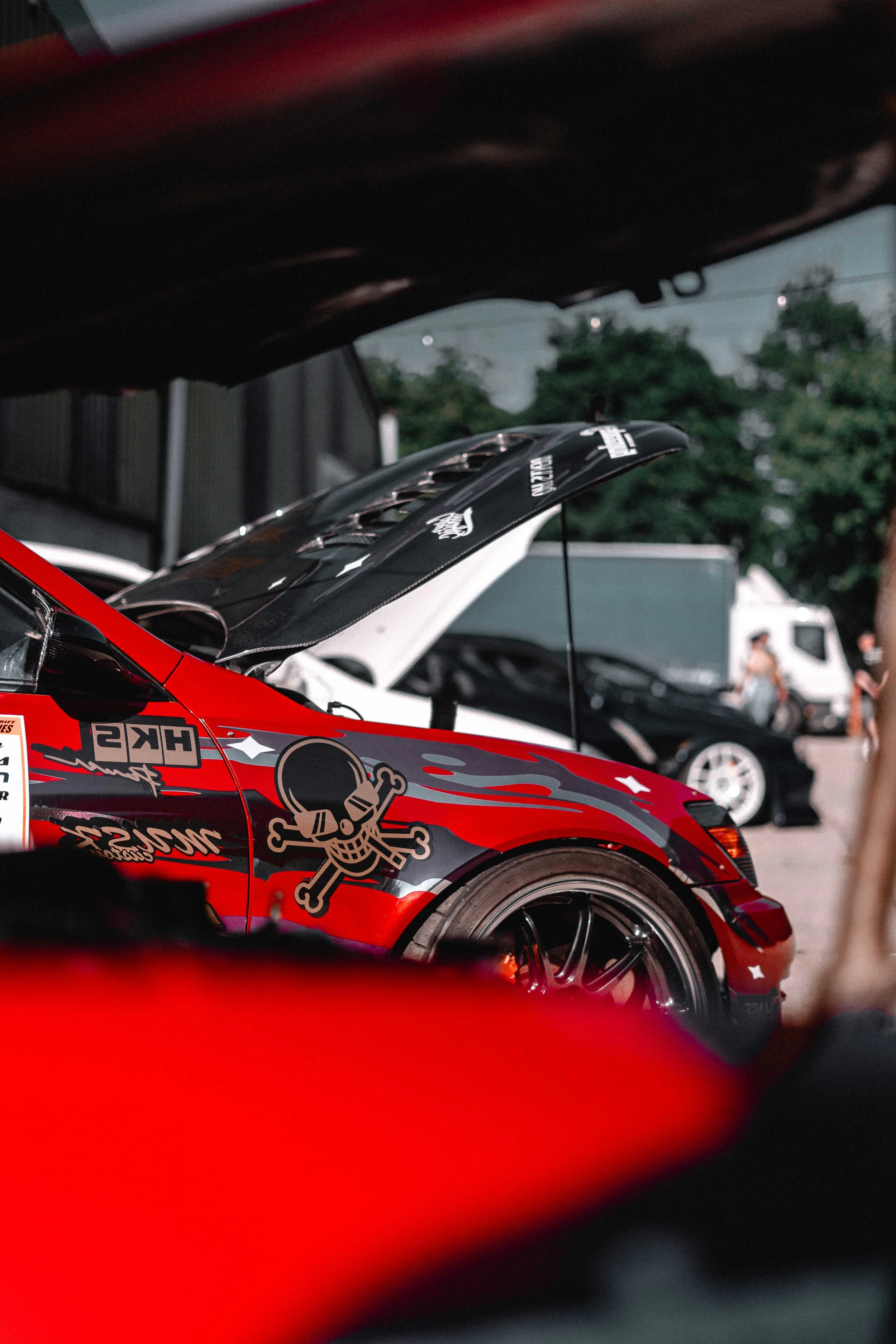 Close-up of a race car with skull and crossbones decal, red and black design, open hood, and black wheel, parked outdoors with other vehicles and people in the background.