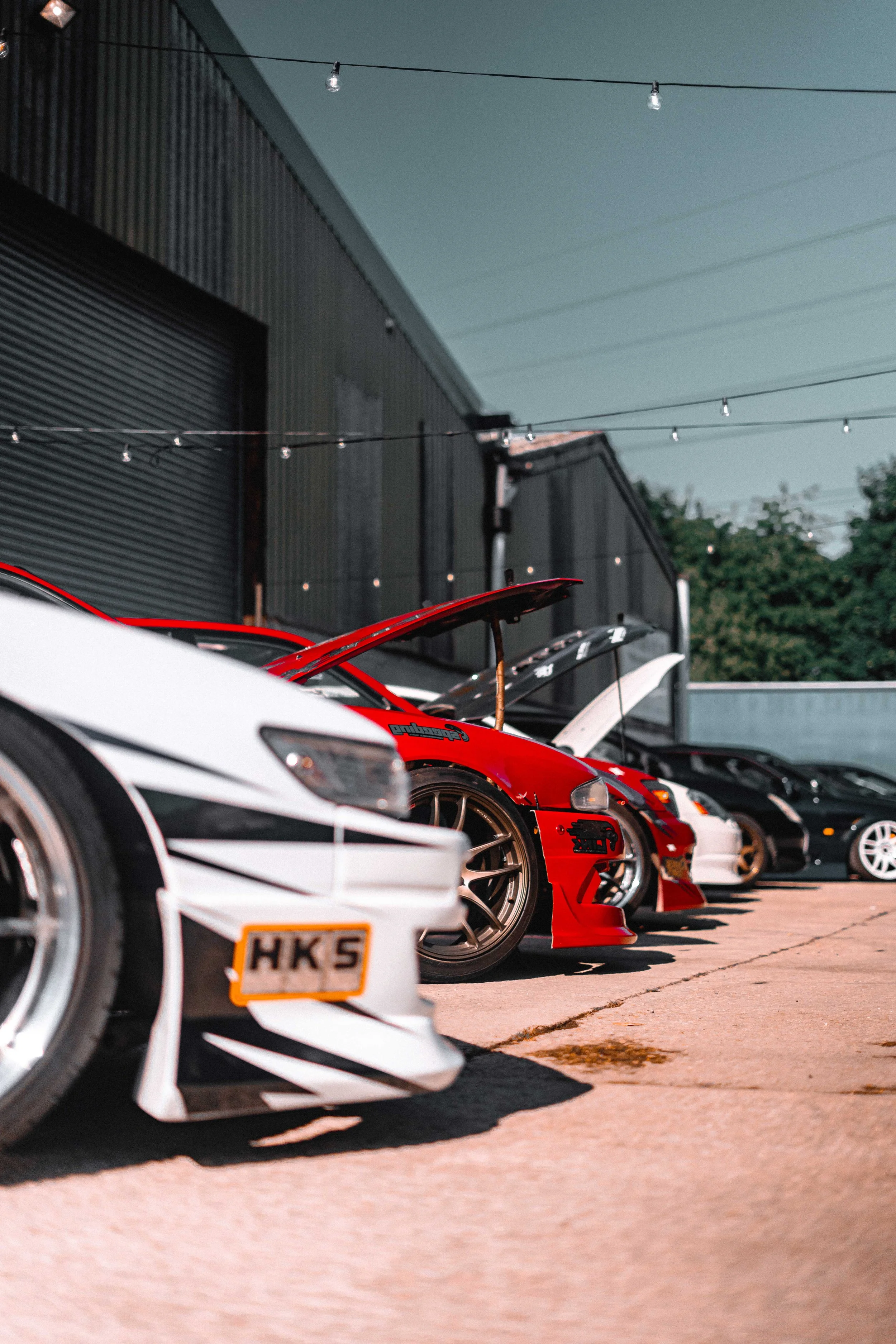 Multiple sports cars parked outside with their hoods open, in a lined-up fashion, under a string of lights, at a car meet or event, during evening hours.
