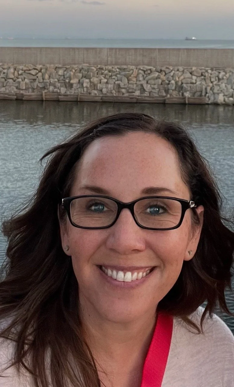 A woman with dark brown hair, wearing glasses and earrings, smiling at the camera, with water, a stone wall, and a cloudy sky in the background.
