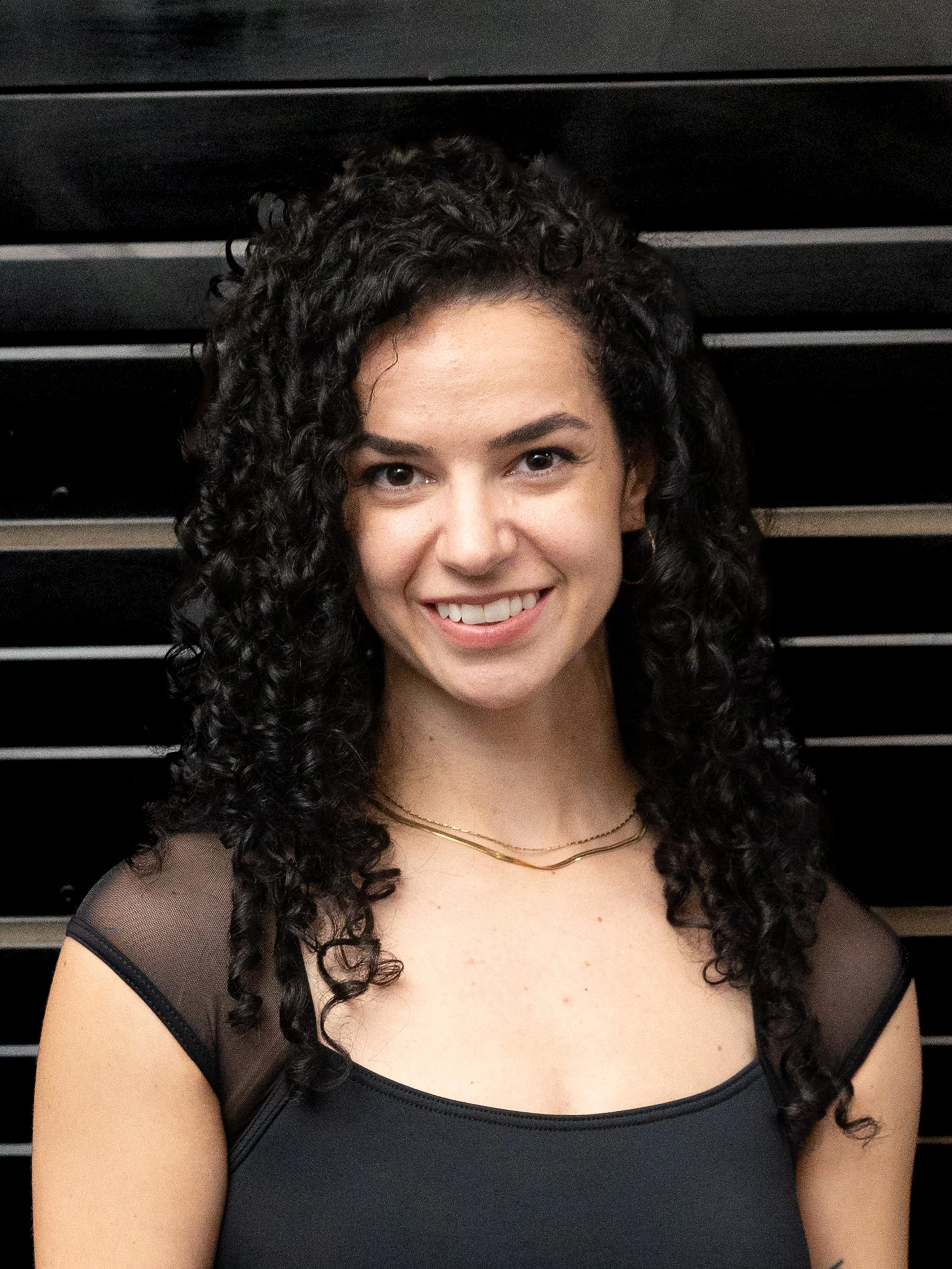 Close-up photograph of a woman with dark curly hair, smiling, wearing a black sheer top with short sleeves, and layered gold necklaces. The background features black horizontal slats.