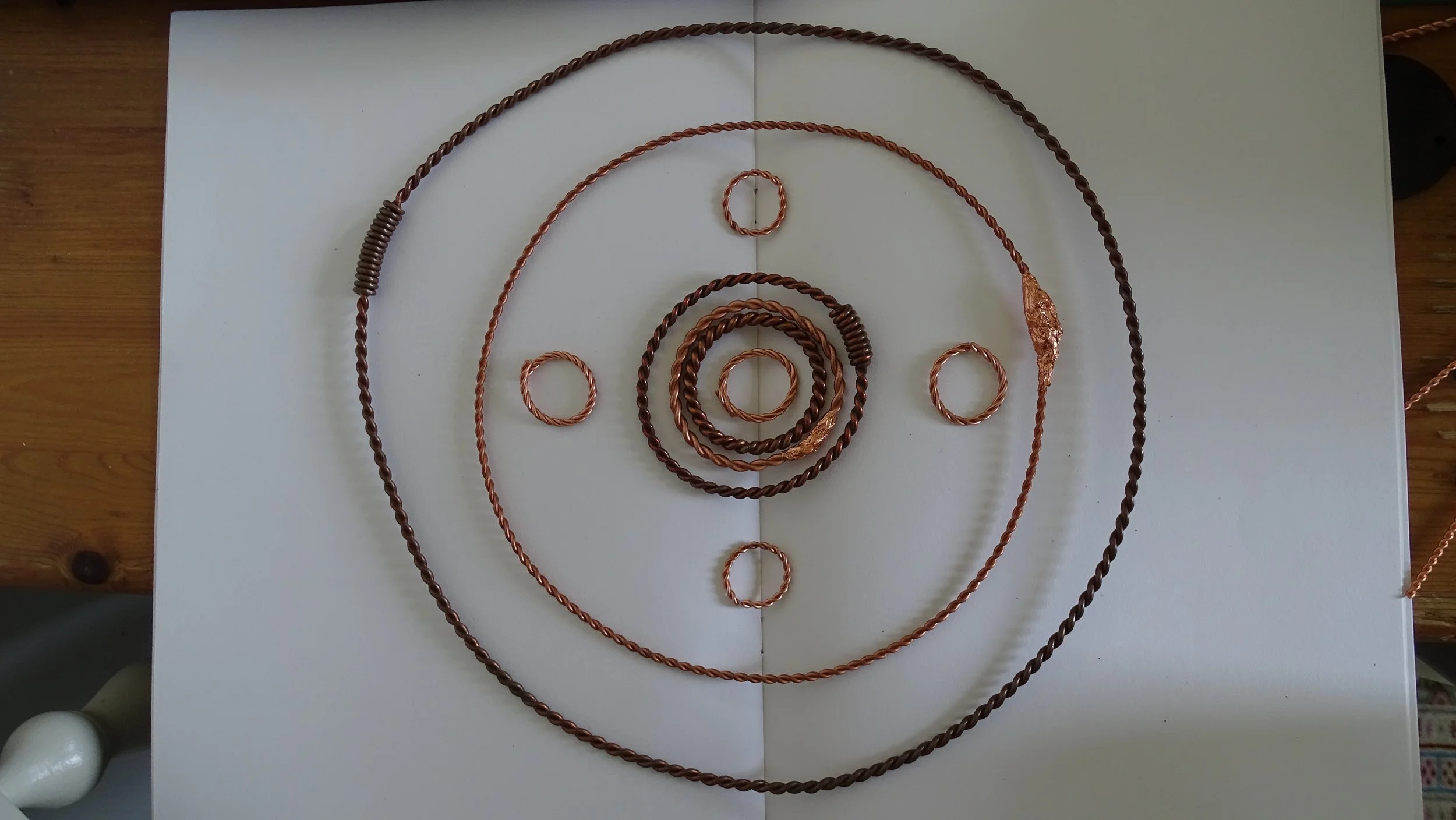 Arrangement of copper wires in a spiral pattern on a white surface. The wires are coiled into different sizes, creating concentric circles.