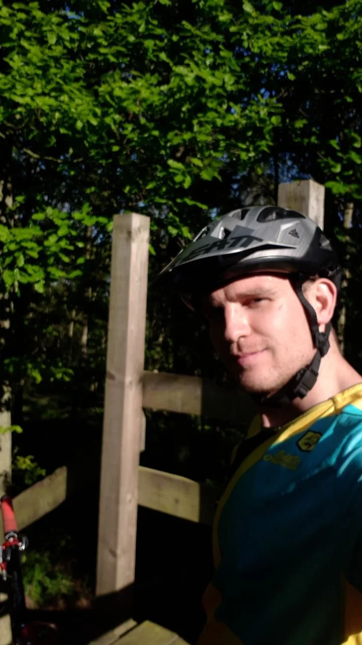 A man wearing a helmet and cycling gear taking a selfie outdoors near a wooden fence and green trees.