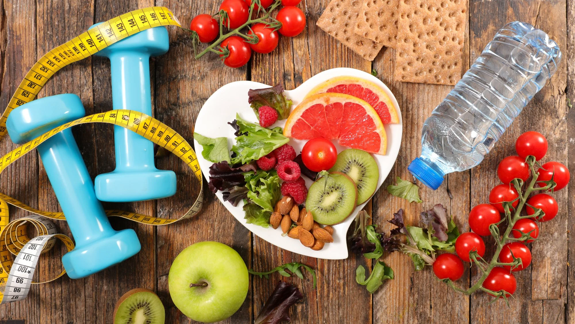 Healthy lifestyle items on a wooden table, including blue dumbbells, a yellow measuring tape, a white heart-shaped plate with mixed fruits and nuts, a green apple, cherry tomatoes, a water bottle, whole grain crackers, and scattered leafy greens.